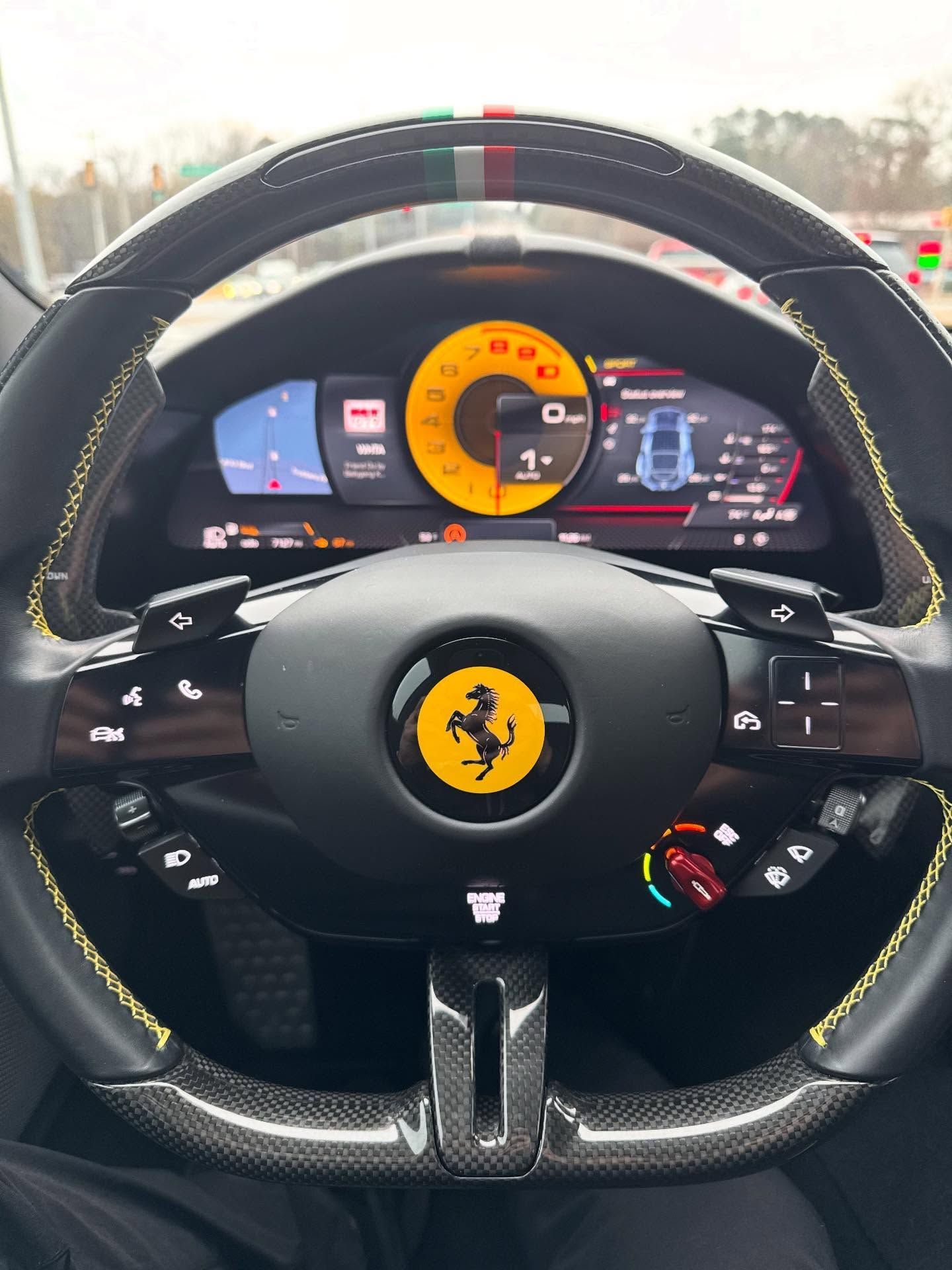 First-person view of a carbon-fiber Ferrari steering wheel with yellow stitching and a digital dashboard on a road.