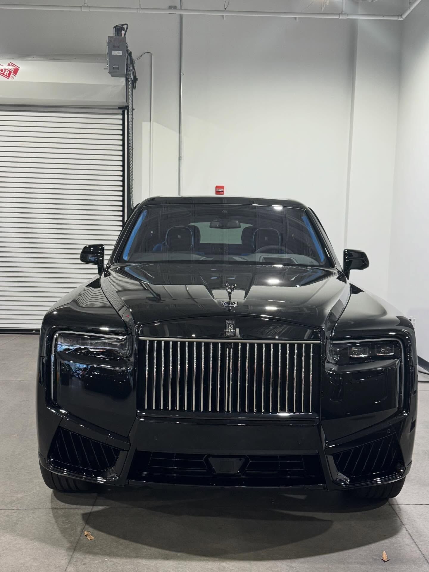 A front view of a black Rolls-Royce sedan parked in a garage with a metallic rolling door in the background.