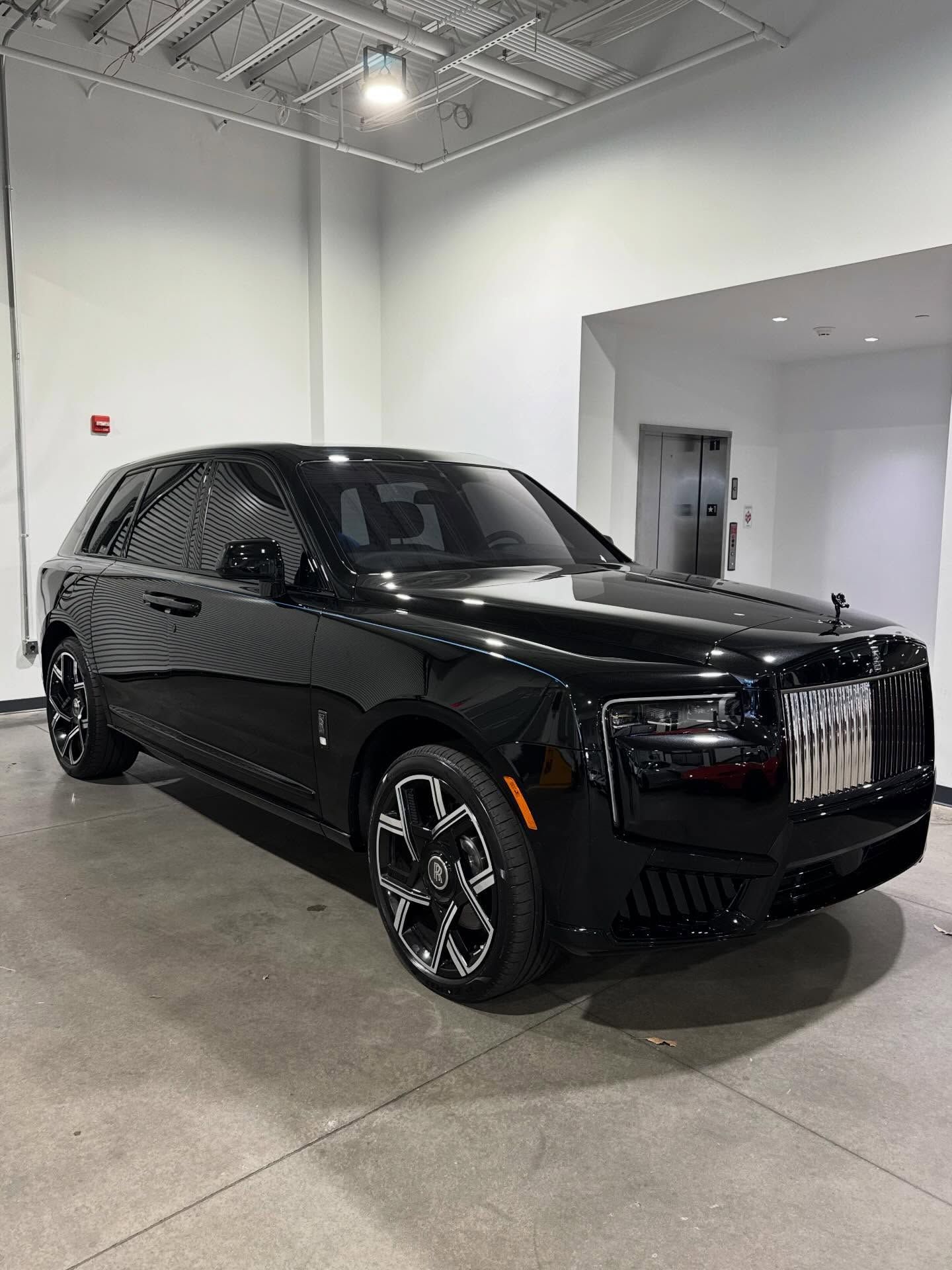 A black Rolls-Royce Cullinan parked indoors on a concrete floor with bright overhead lighting.