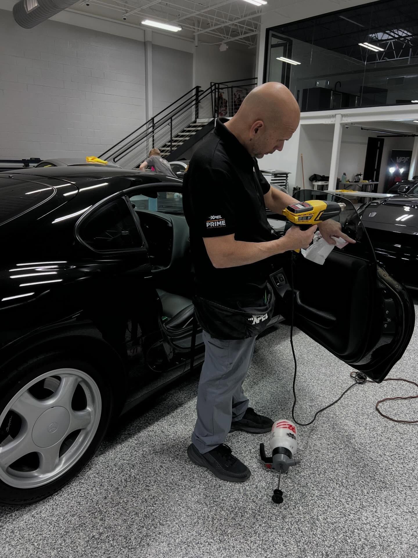 A person uses a yellow tool to work on the interior of a black car in a garage with a staircase in the background.