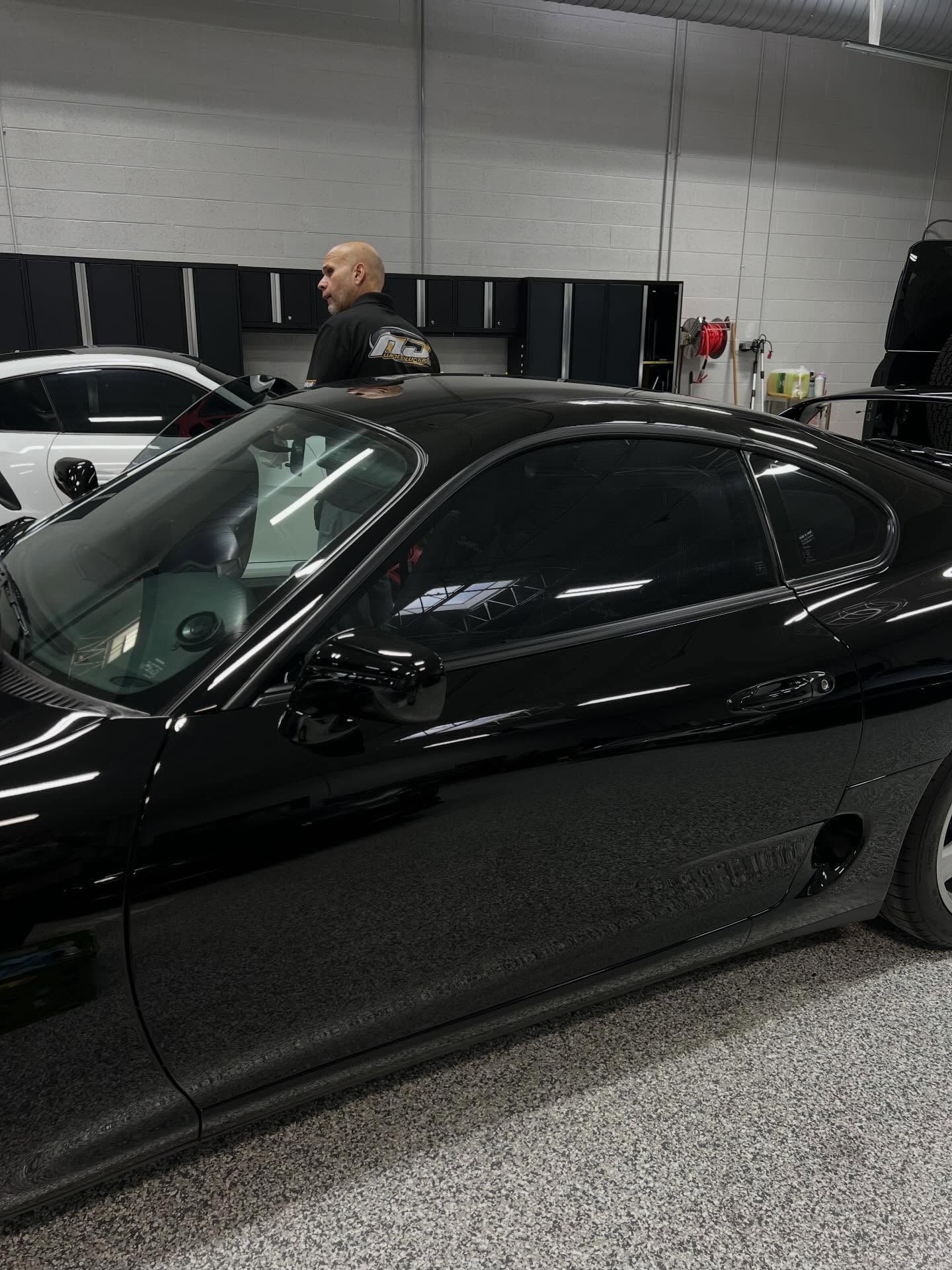A bald person standing in a garage beside a black Toyota Supra, with another car partially visible in the background.