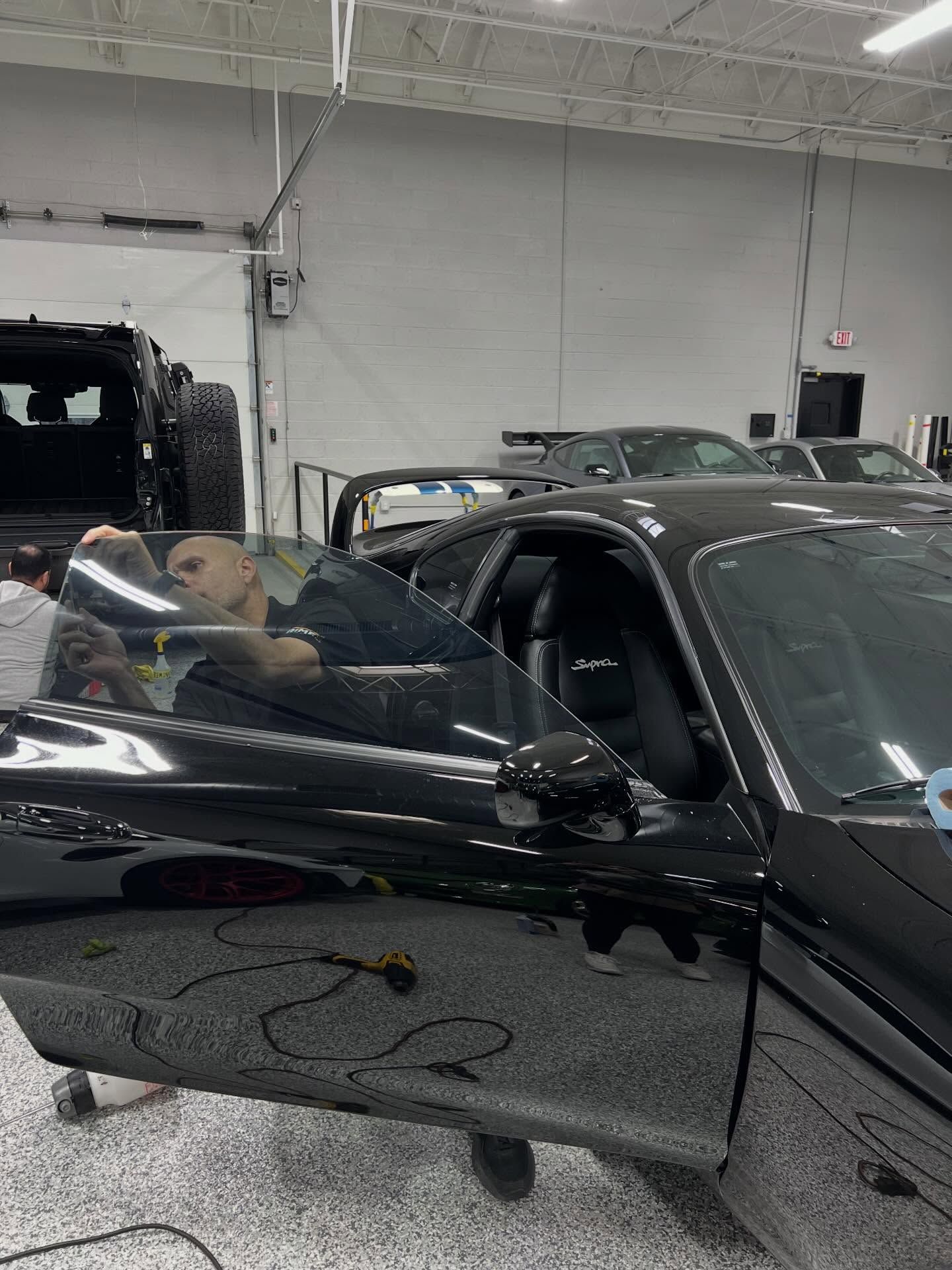 A person inside a garage is applying window tint to the driver-side window of a black sports car.