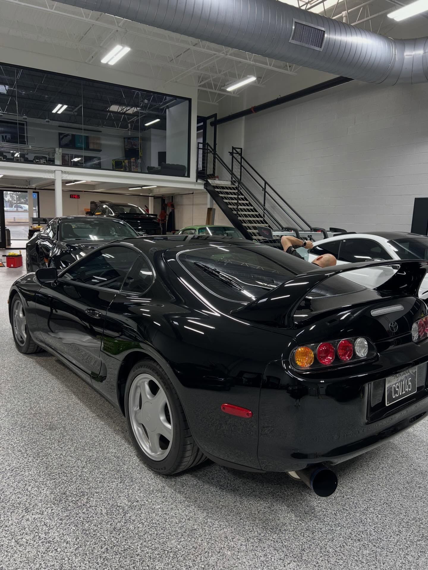 A black Toyota Supra parked in a garage with a staircase and mezzanine level in the background.