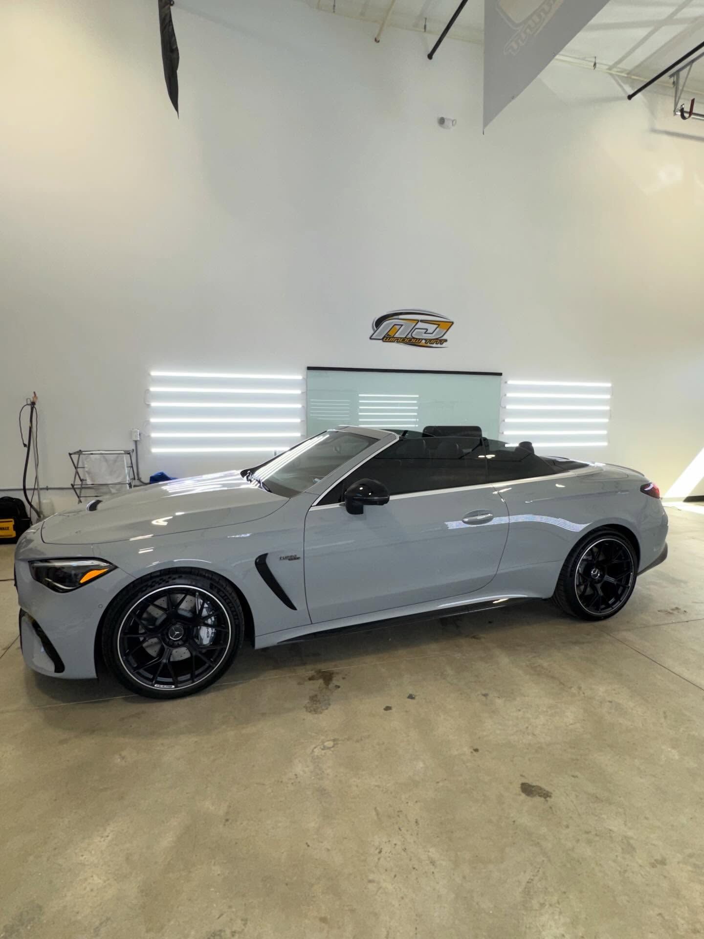 A light grey Mercedes-AMG convertible parked in a brightly lit, professional garage with white walls and a logo.