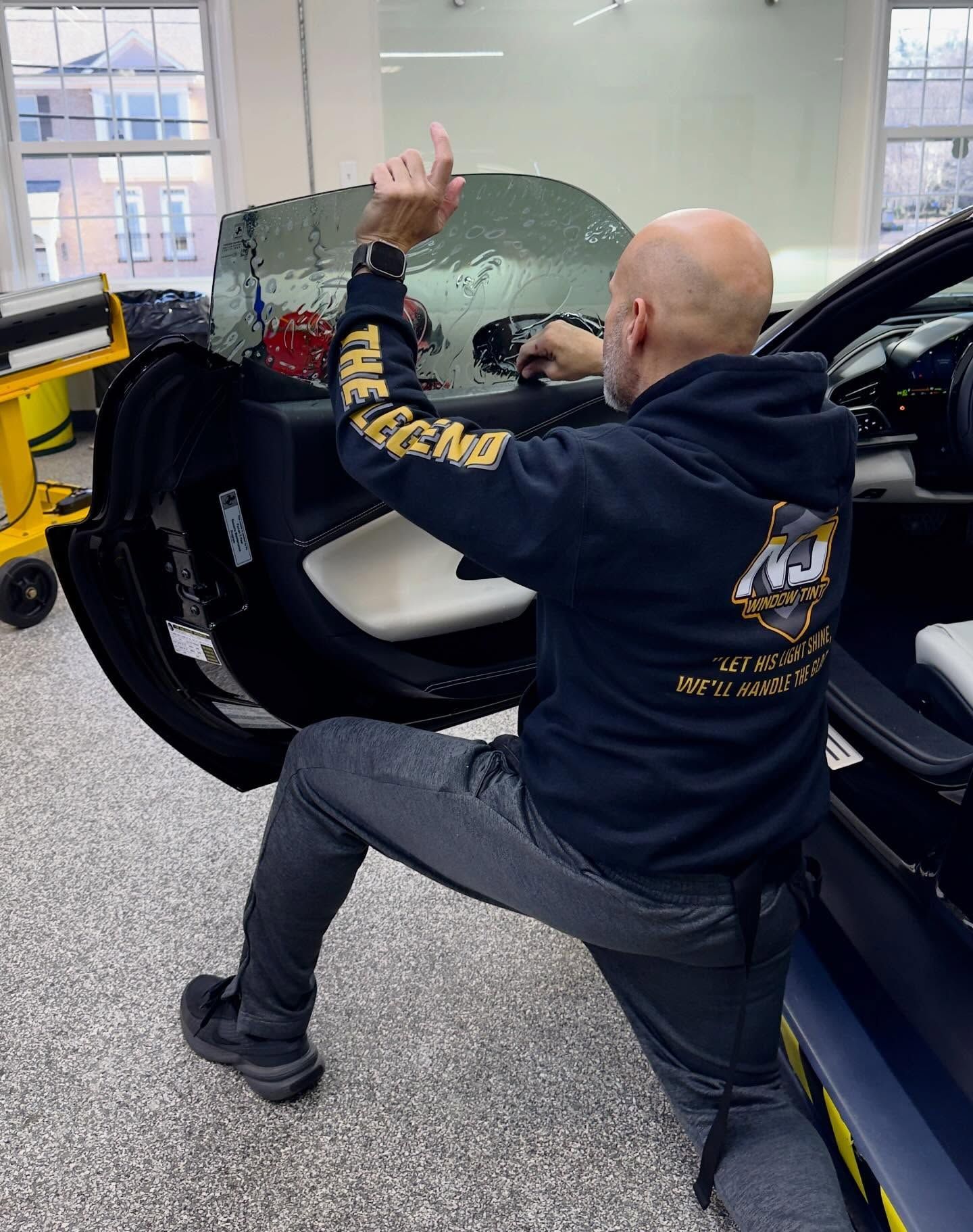 A technician in a black hoodie applies window tint to a car door inside a shop.