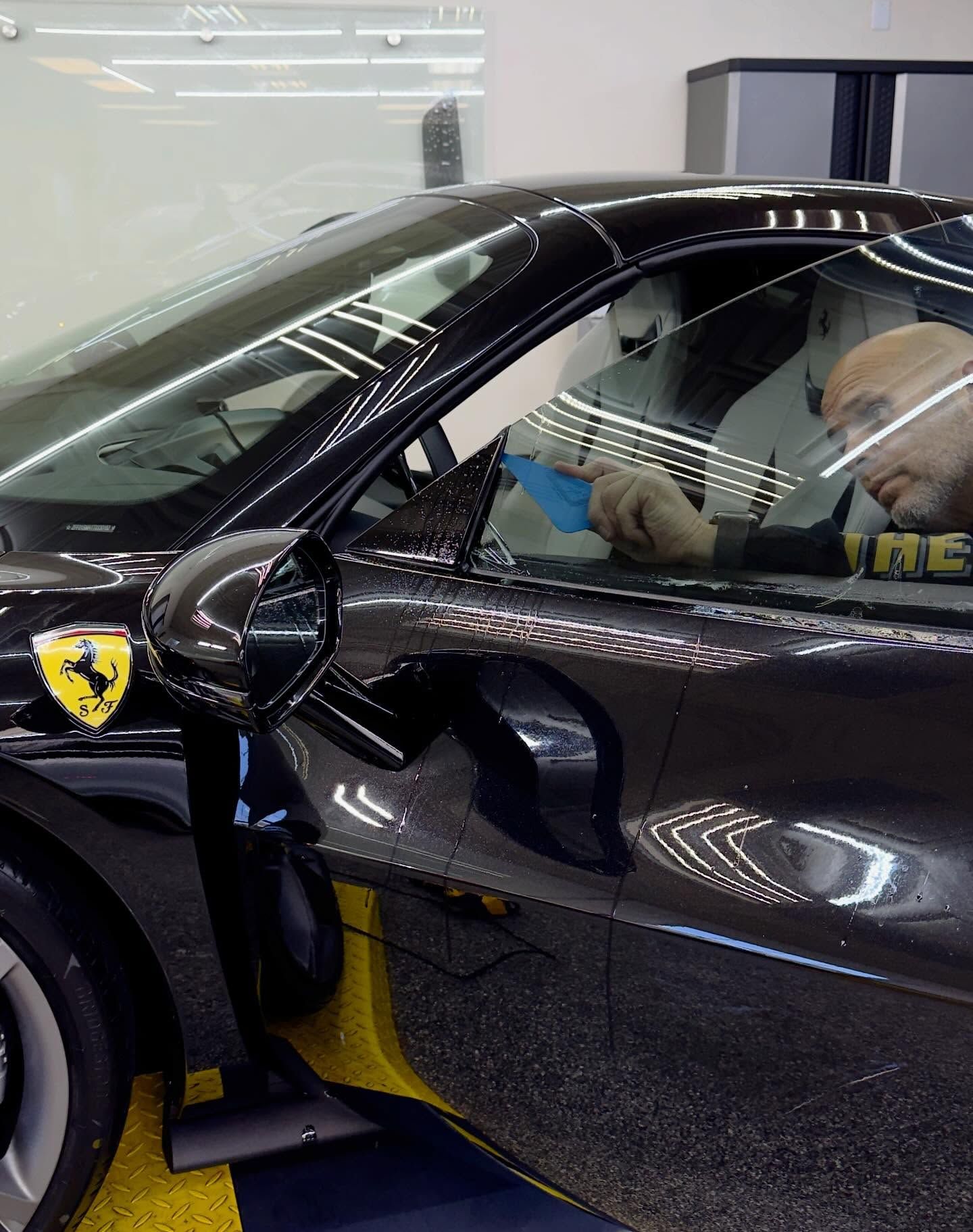 A technician carefully applies window tint to the driver-side door of a black Ferrari in a professional workshop.