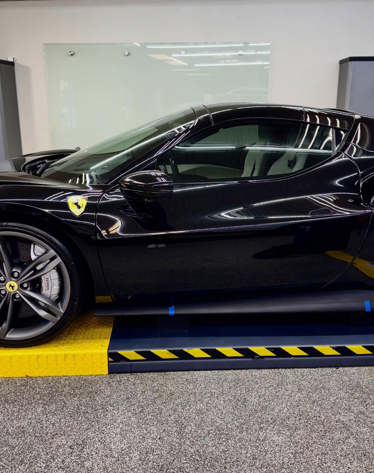 A side view of a glossy black Ferrari sports car parked on a yellow and black industrial service lift in a showroom.