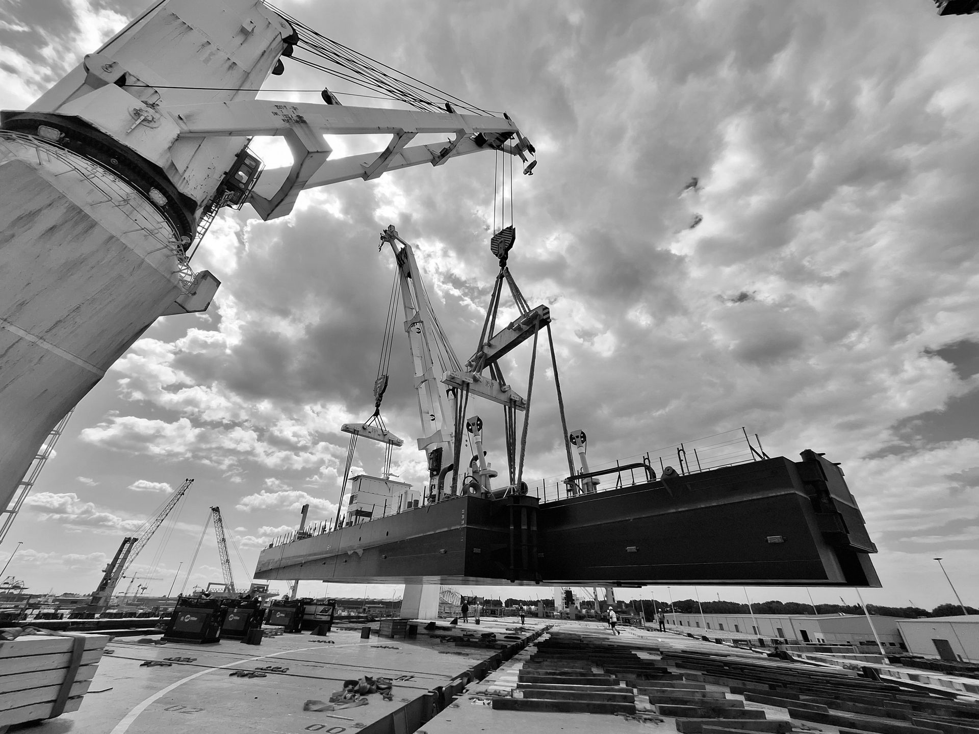 Een component van een vrachtschip wordt door kranen opgetild in een haven, onder een bewolkte hemel.