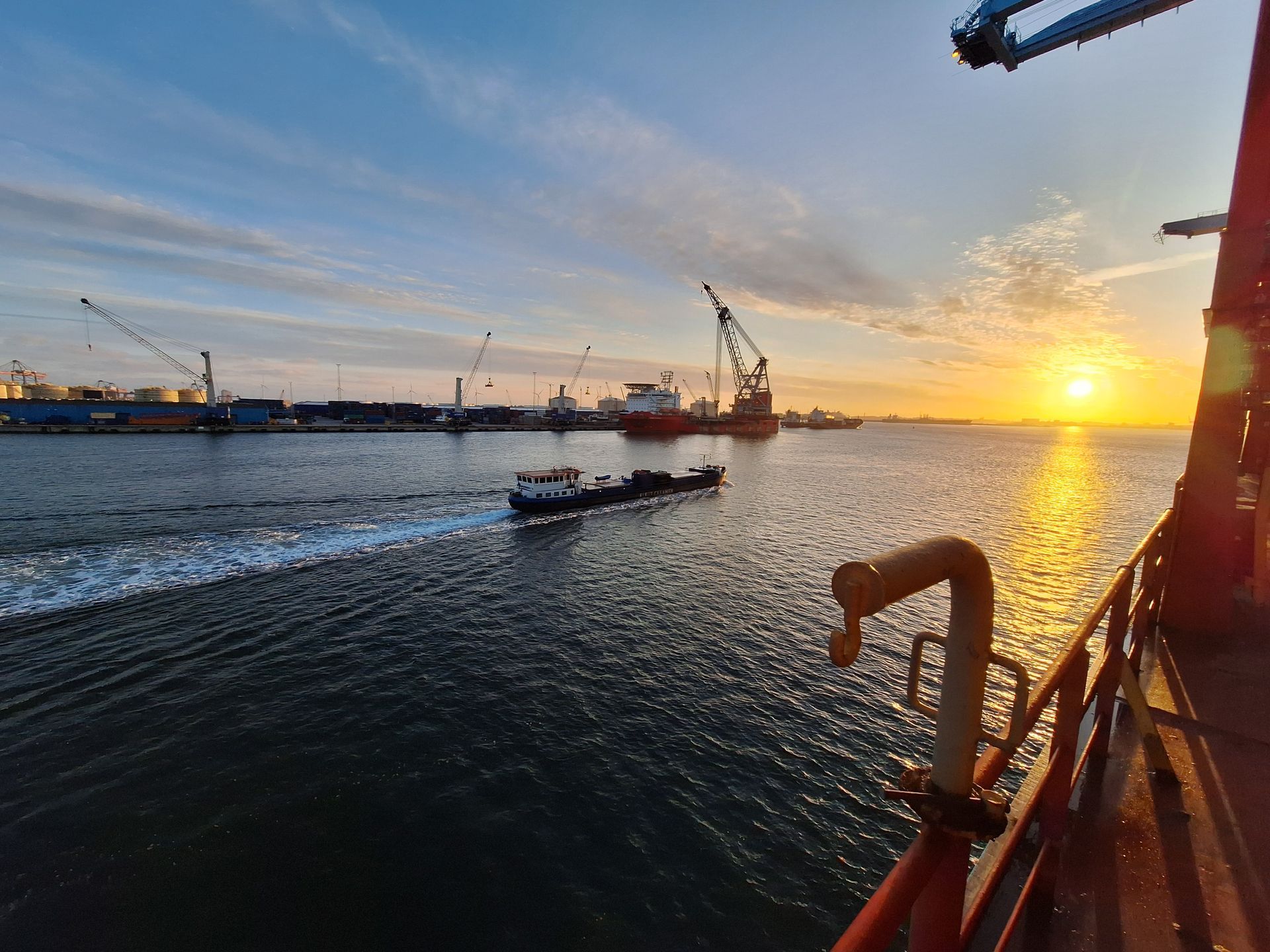 Sunrise over harbor, small boat traveling, cranes and ships at dock, orange and blue sky. P&I en maritieme partijen support -