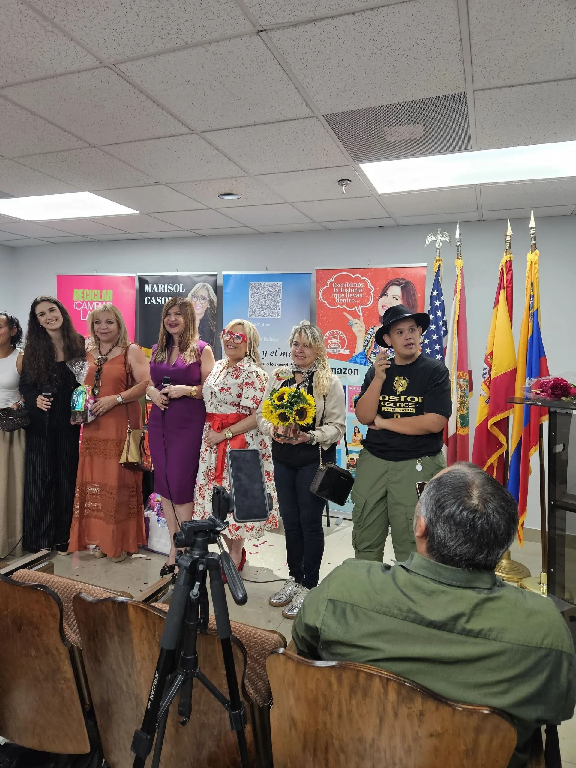 People on a stage at an indoor event, with flags, banners, and cameras in the foreground