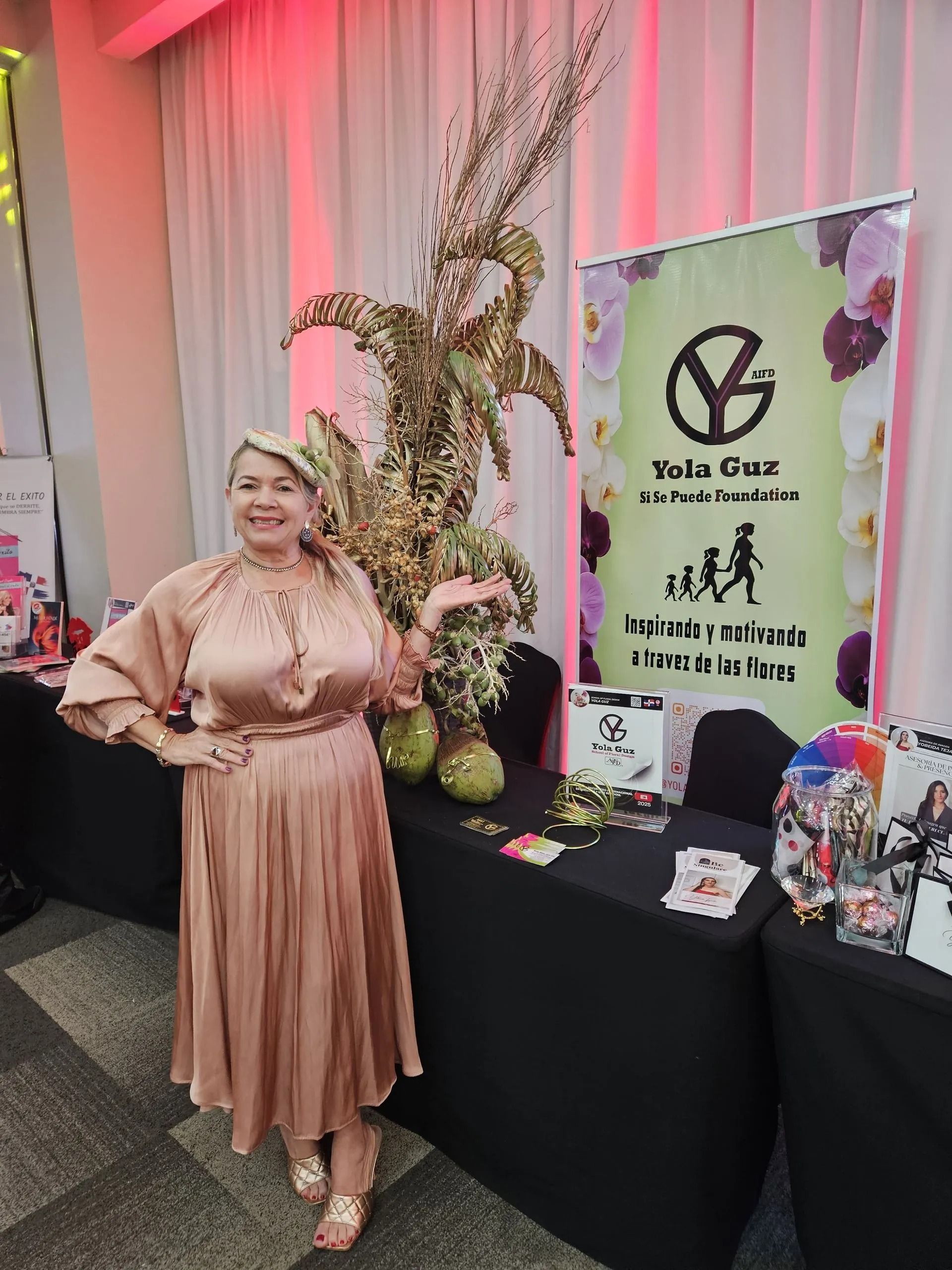 Woman in a pink dress posing beside a table and event banner with floral decor.