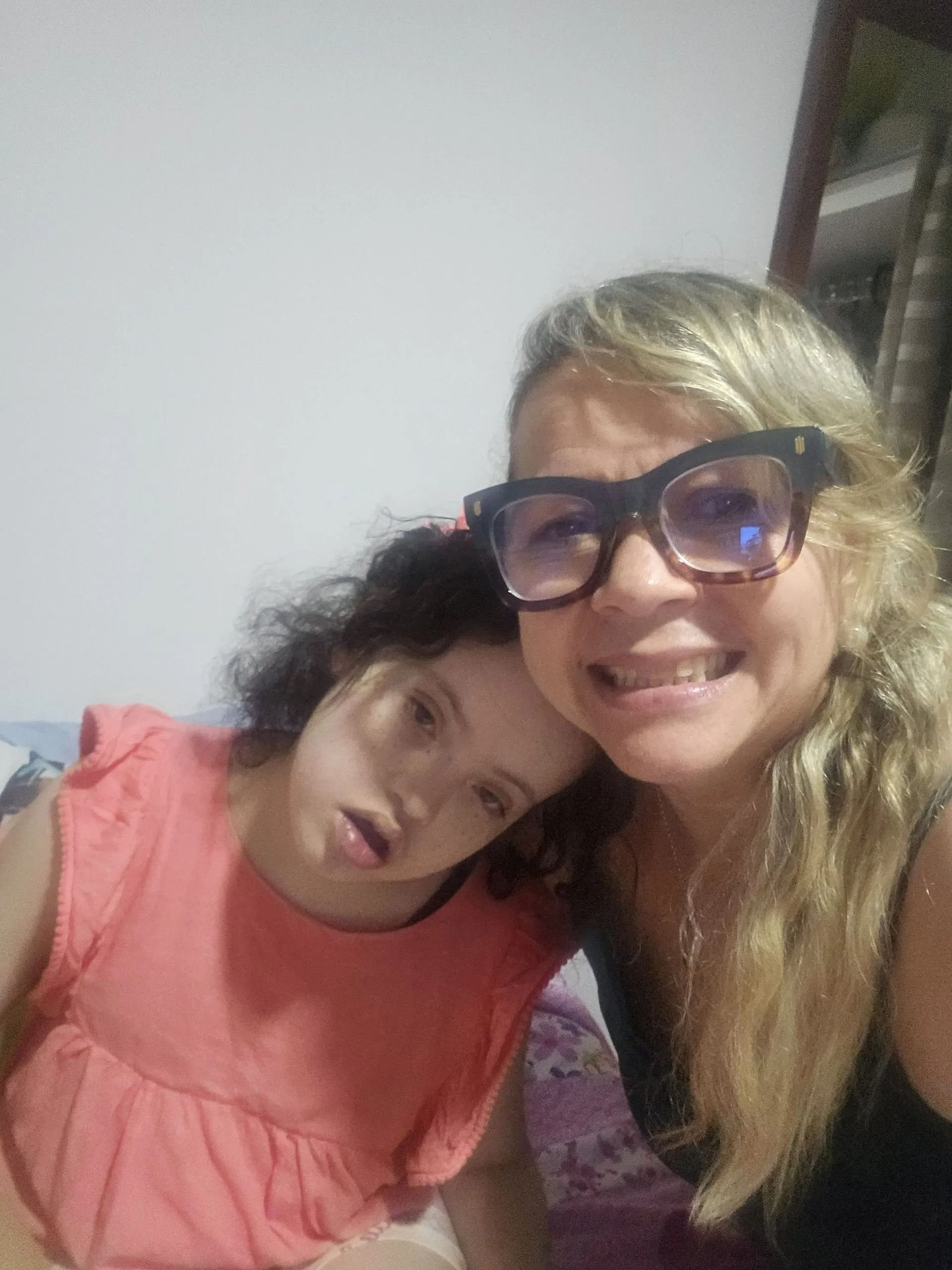 Two smiling people posing closely together indoors, one in a coral top and one in black glasses.