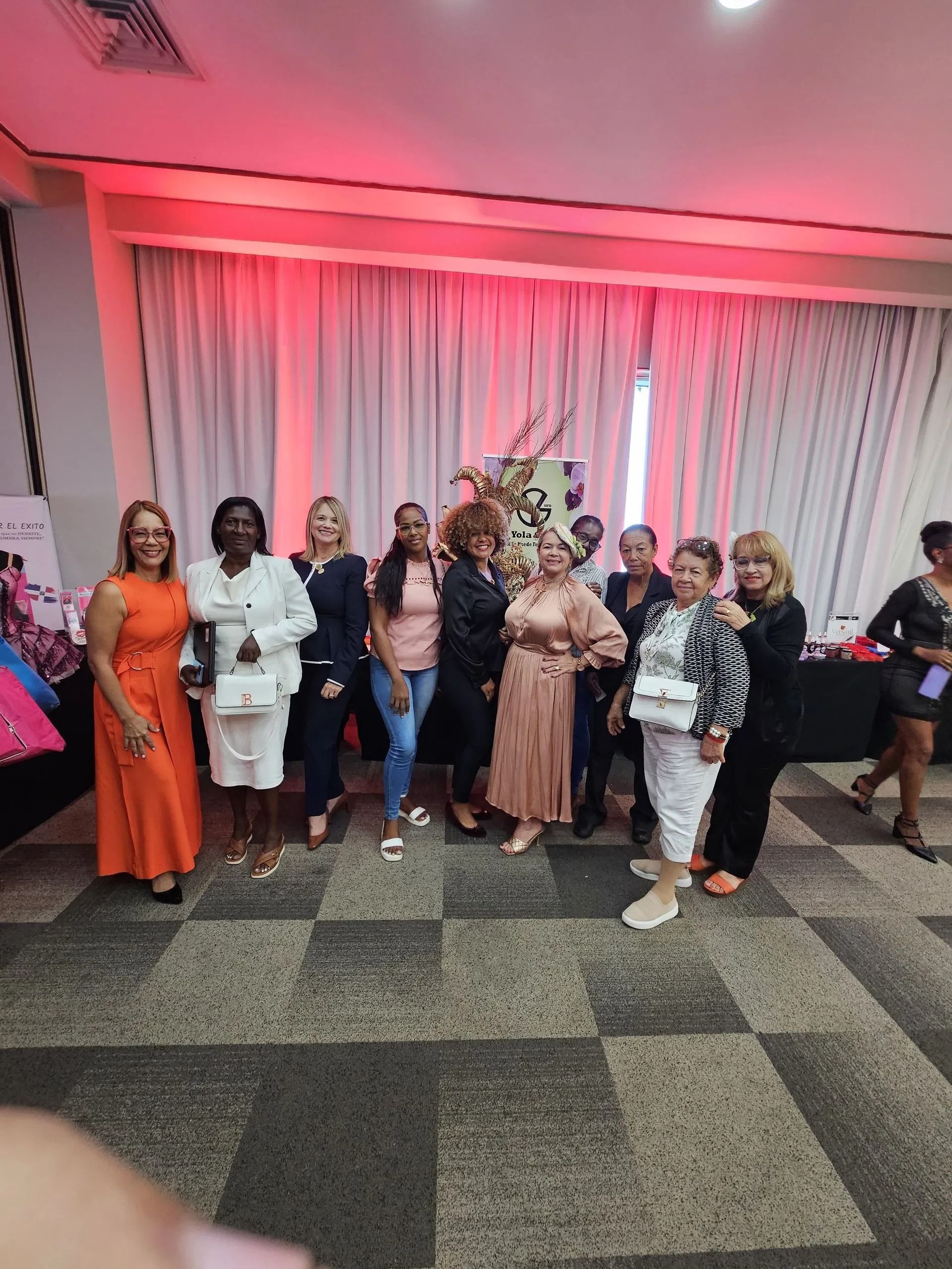 Group of people posing indoors at an event with pink lighting and white drapes backdrop.
