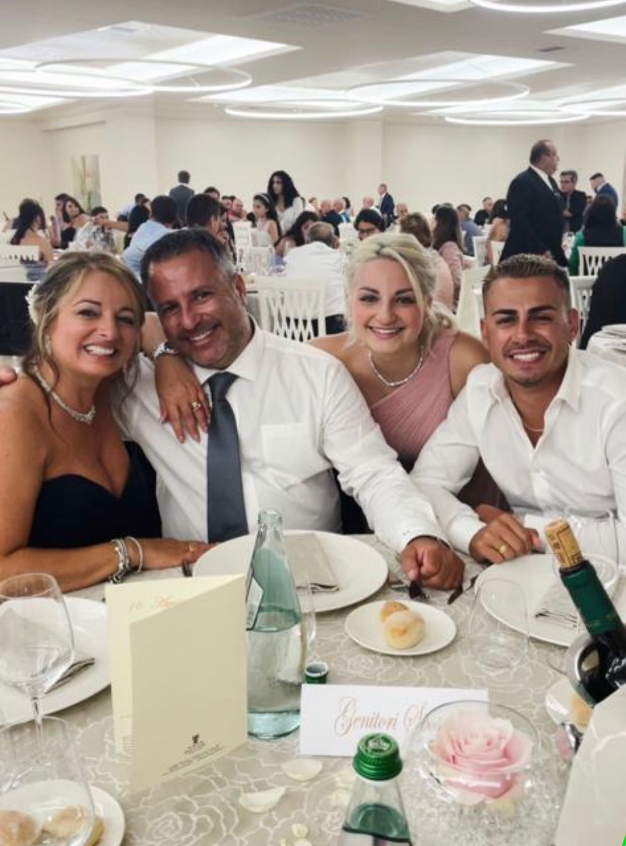 Quattro persone sorridenti a un tavolo apparecchiato per un evento formale. Ambiente interno con altri ospiti sullo sfondo.