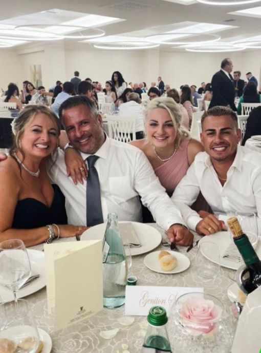 Quattro persone sorridenti a un tavolo apparecchiato per un evento formale. Ambiente interno con altri ospiti sullo sfondo.