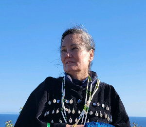 Woman in dark coat with shell necklace, looking out at ocean and sky.