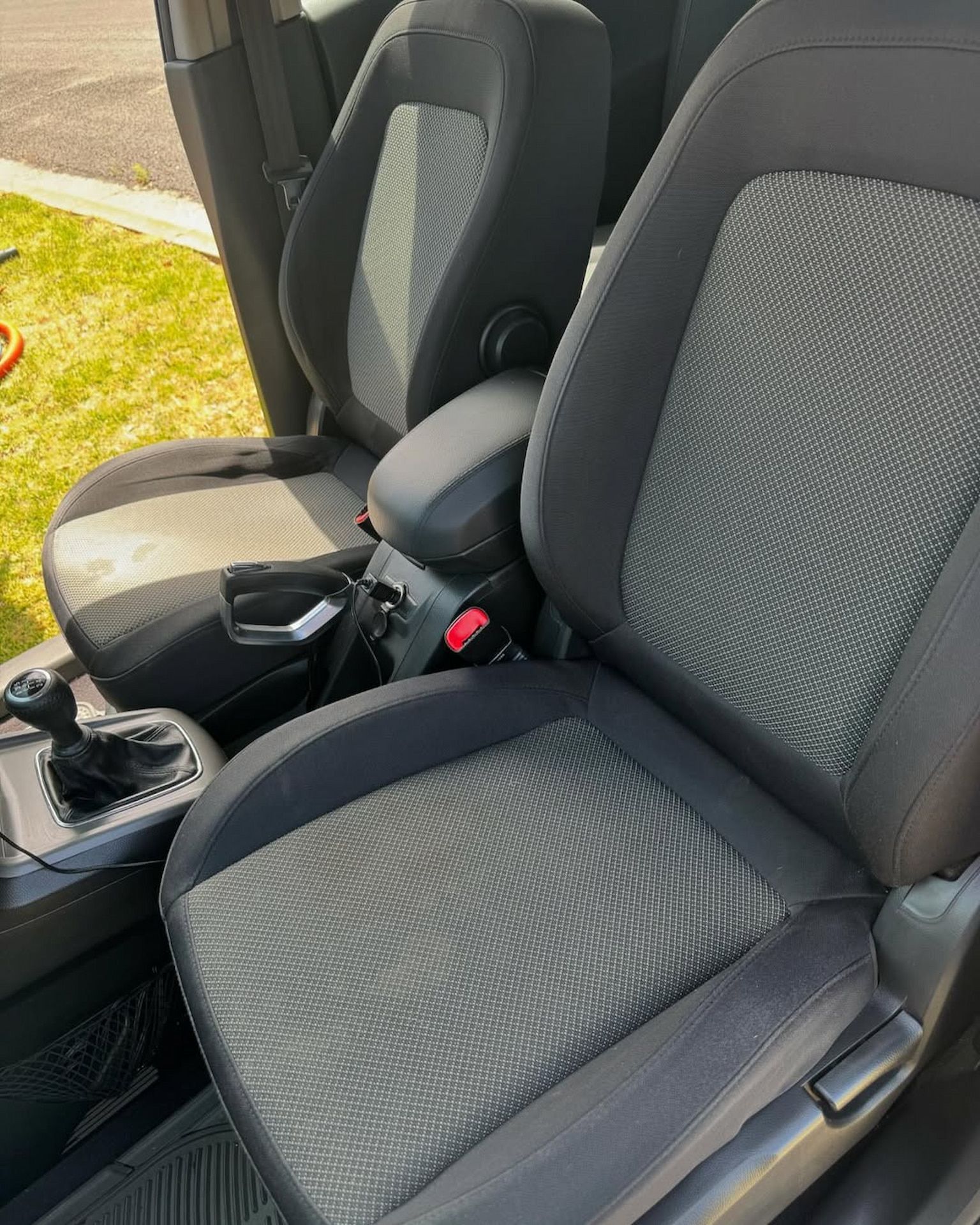 The front interior of a vehicle showing two grey and black patterned fabric bucket seats, a center console, and shifter.