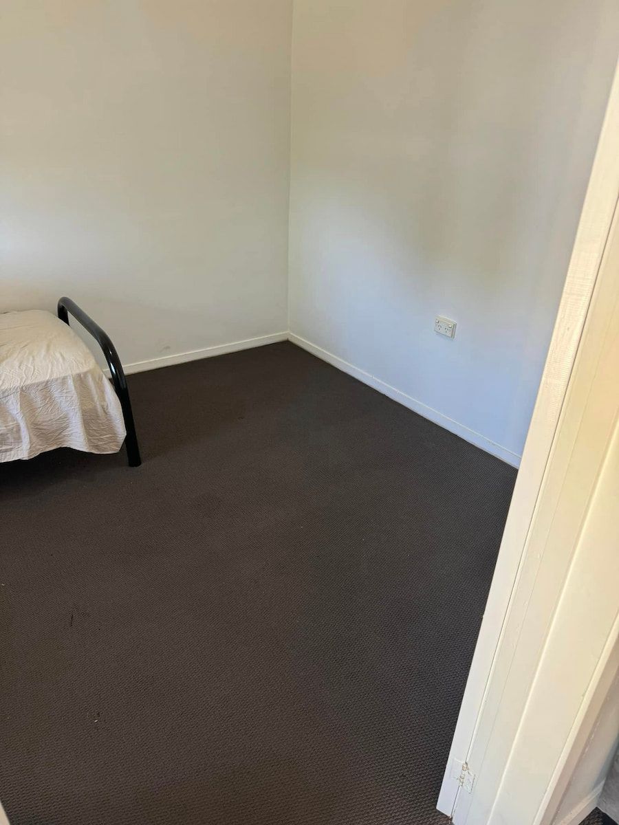 An Empty Room With Grey Carpet, White Walls, and a Single Bed Frame — Cobra's Carpet Cleaning in Junee, NSW