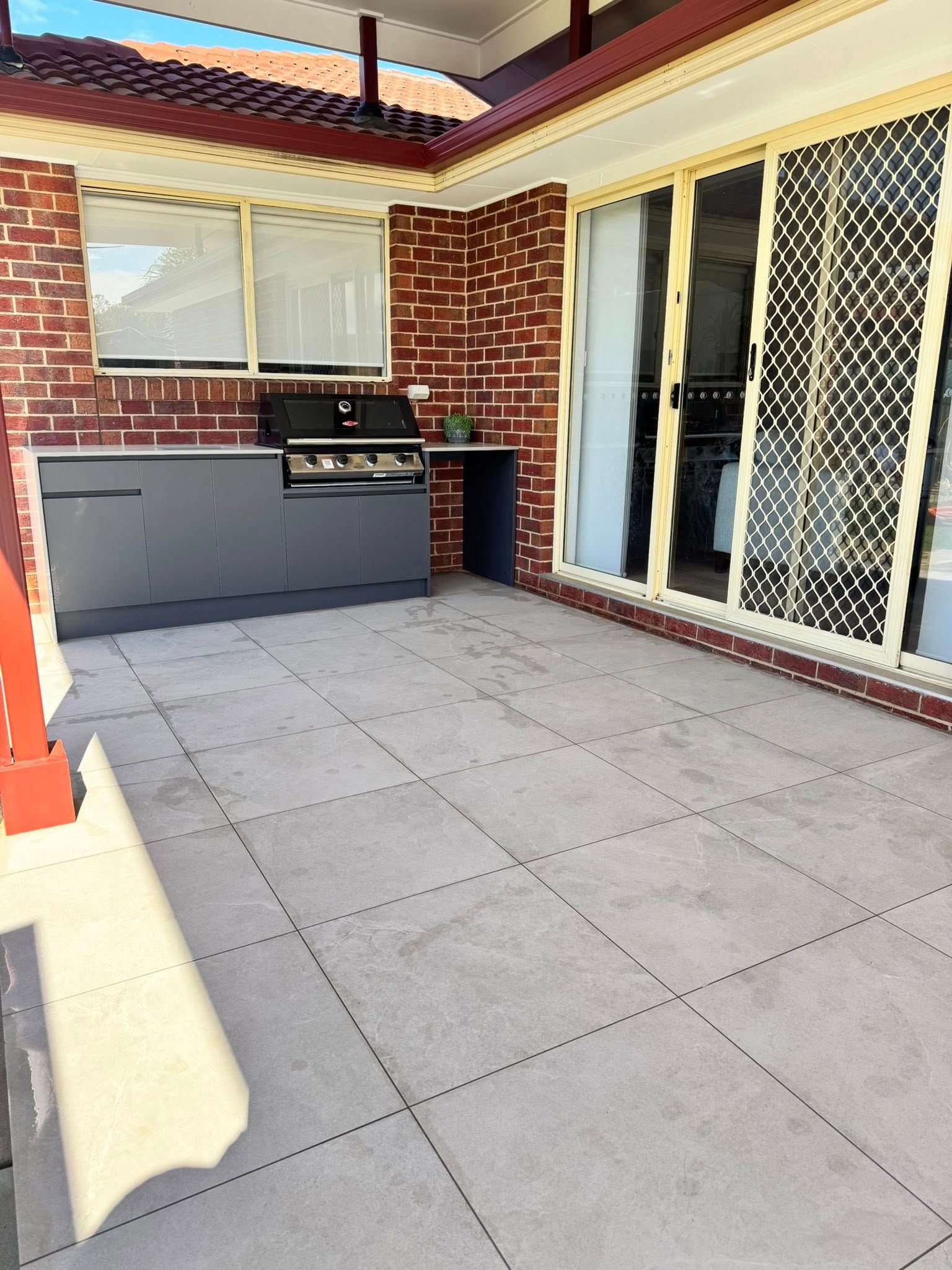 An outdoor patio with light grey stone tiles, a dark grey outdoor kitchen unit with a grill, and sliding glass doors   — Cobra's Carpet Cleaning in Junee, NSW
