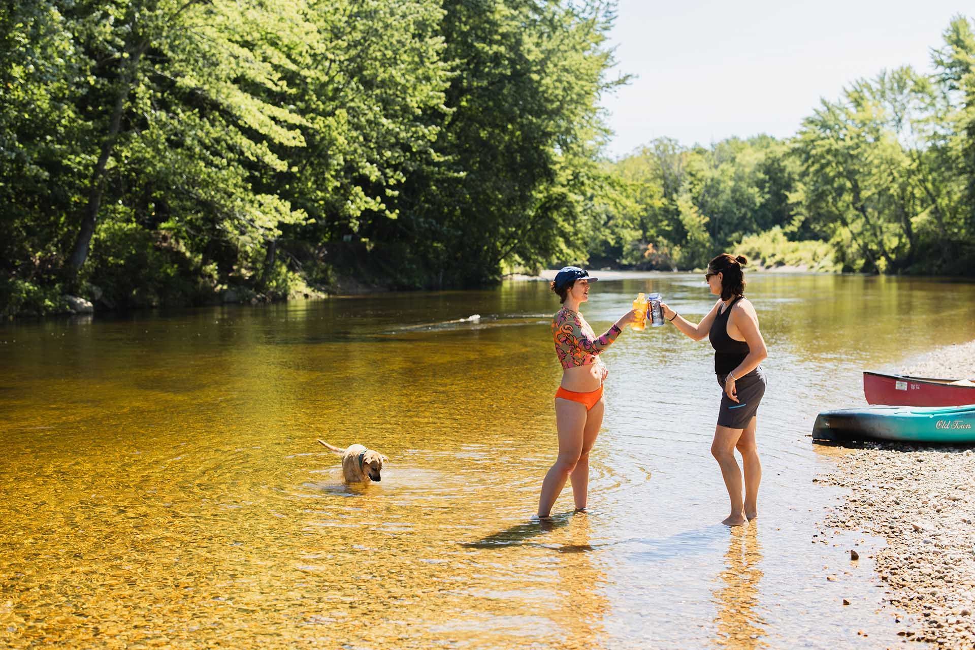 two woman drinking while in the saco river with a dog