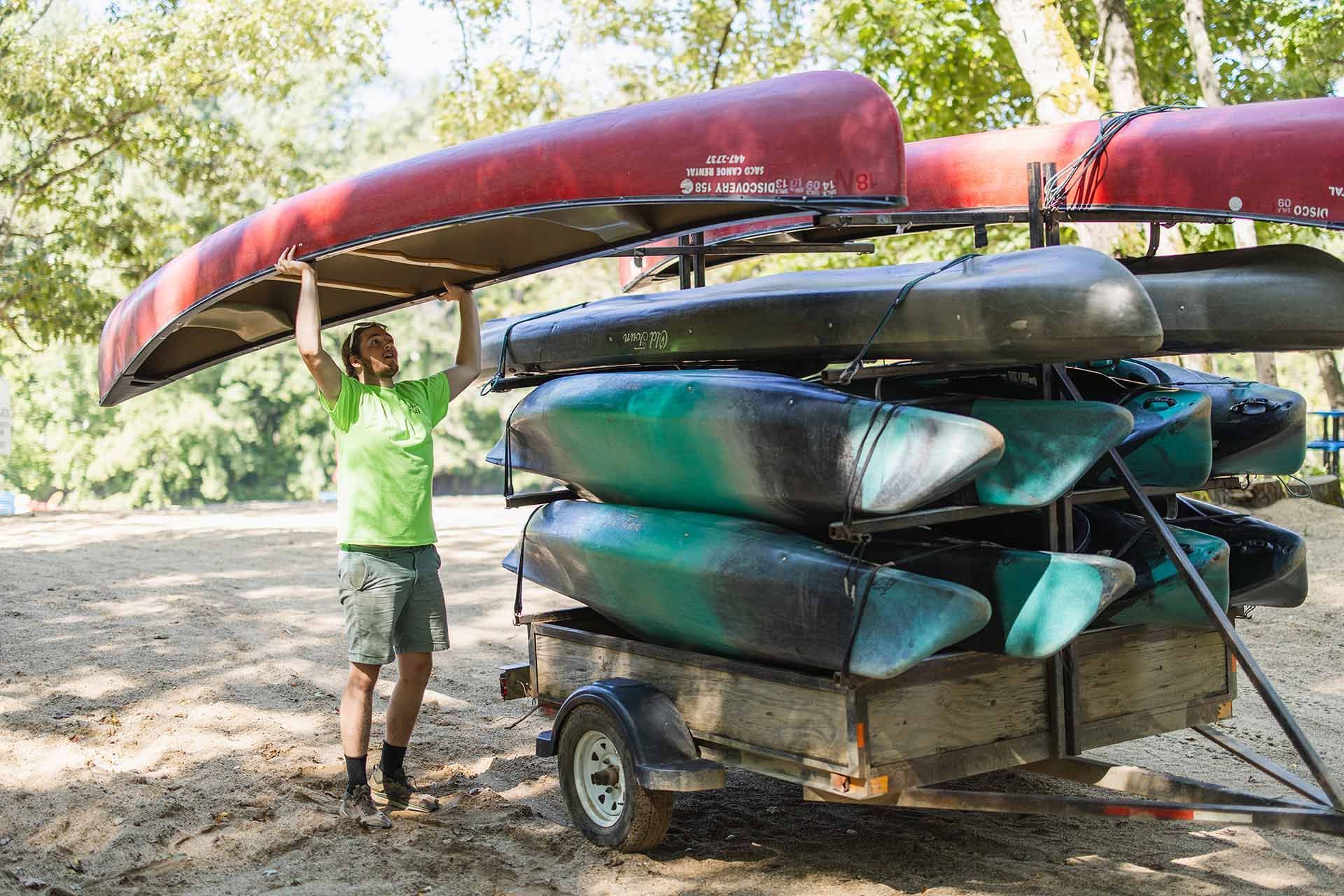 Man Carrying a Canoe Over a Trailer Filled with Kayaks