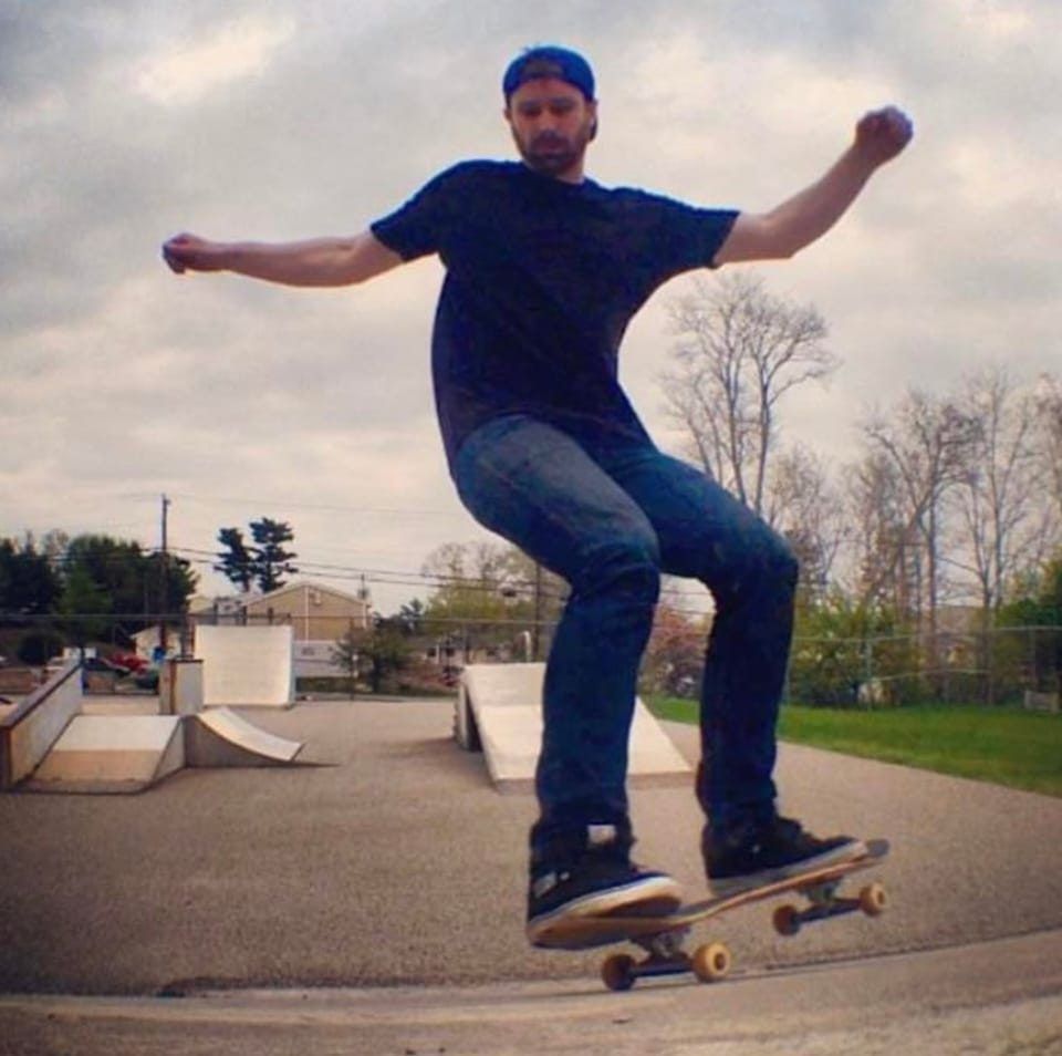 Skateboarder balancing on a board at a skate park, arms outstretched, with ramps in the background.