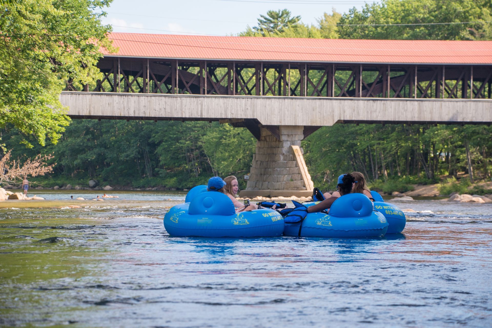 children on river tubes going down the saco river under a bridge