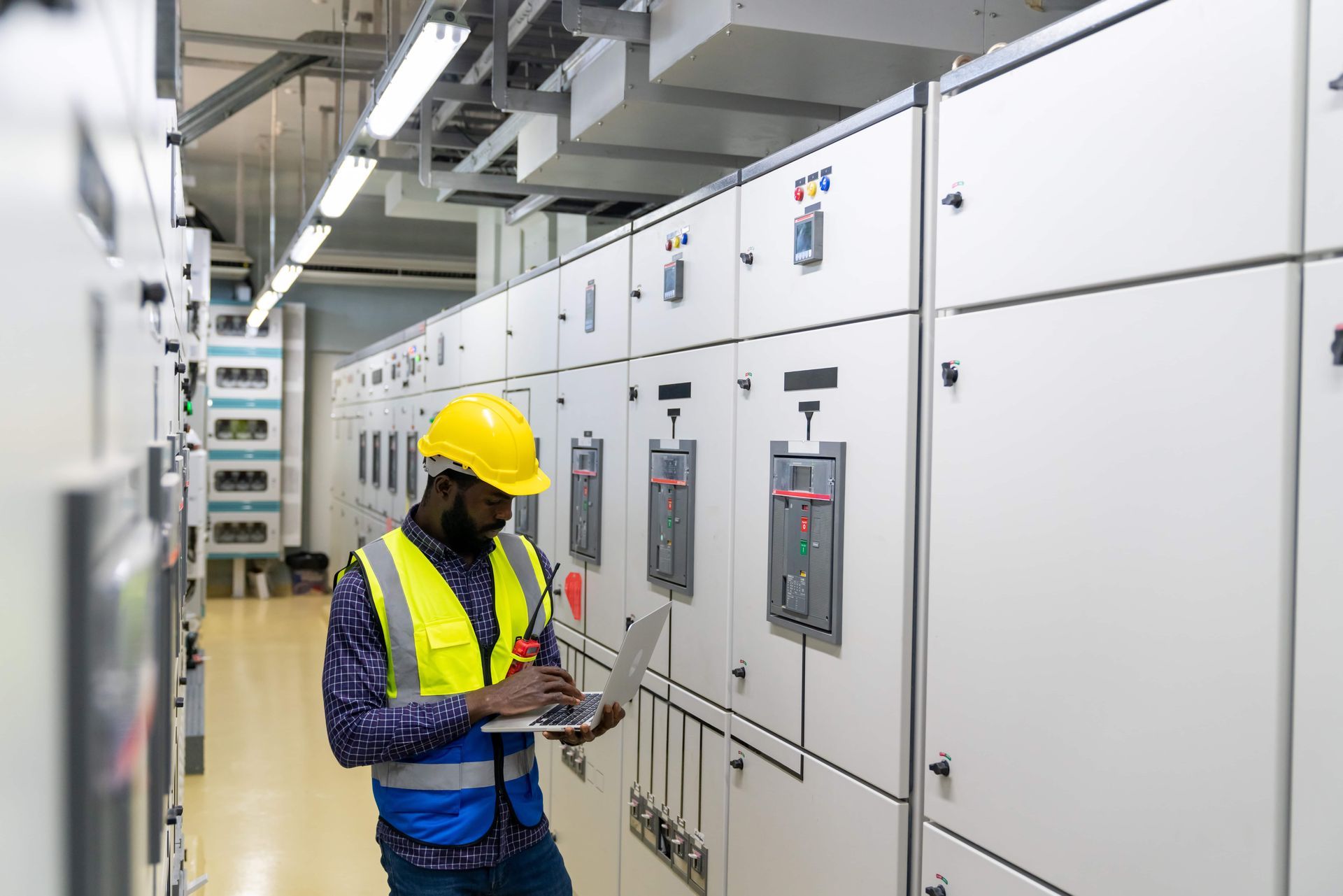 Professional African male engineer in safety uniform working at factory server electric control panel room.