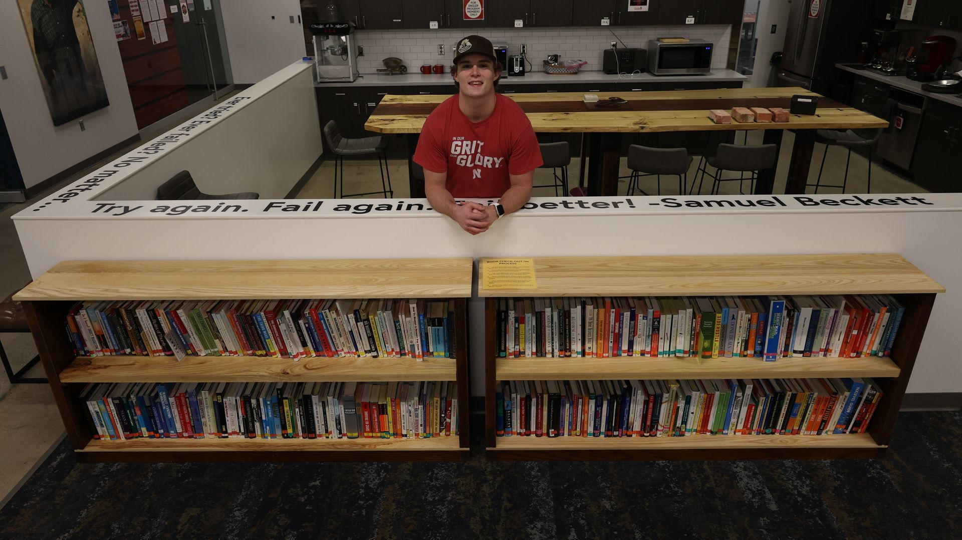 A person stands behind a bookshelf with books, wearing a red shirt and hat. In a library-like space.