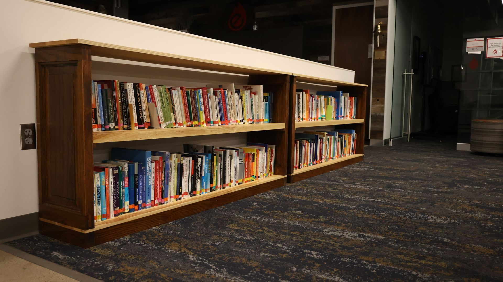 Bookshelves filled with books in a library, dark wood frame, light top. Dark carpeted floor.