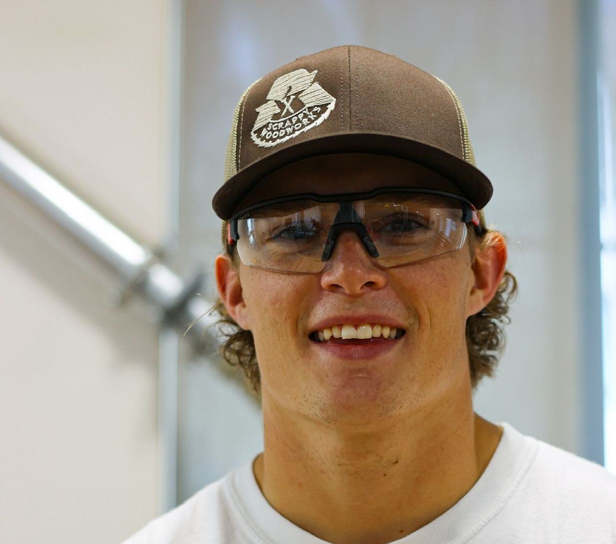 Young man in a brown hat and safety glasses smiles in an indoor setting.