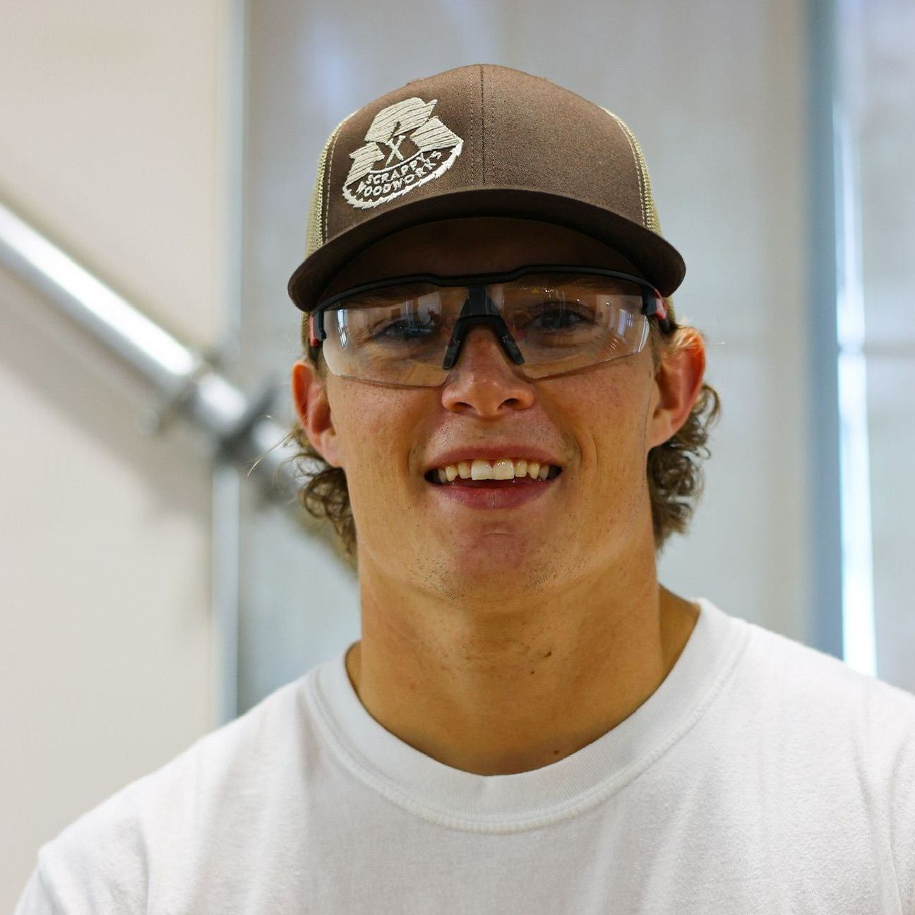 Young man with brown hat and glasses smiling, inside.