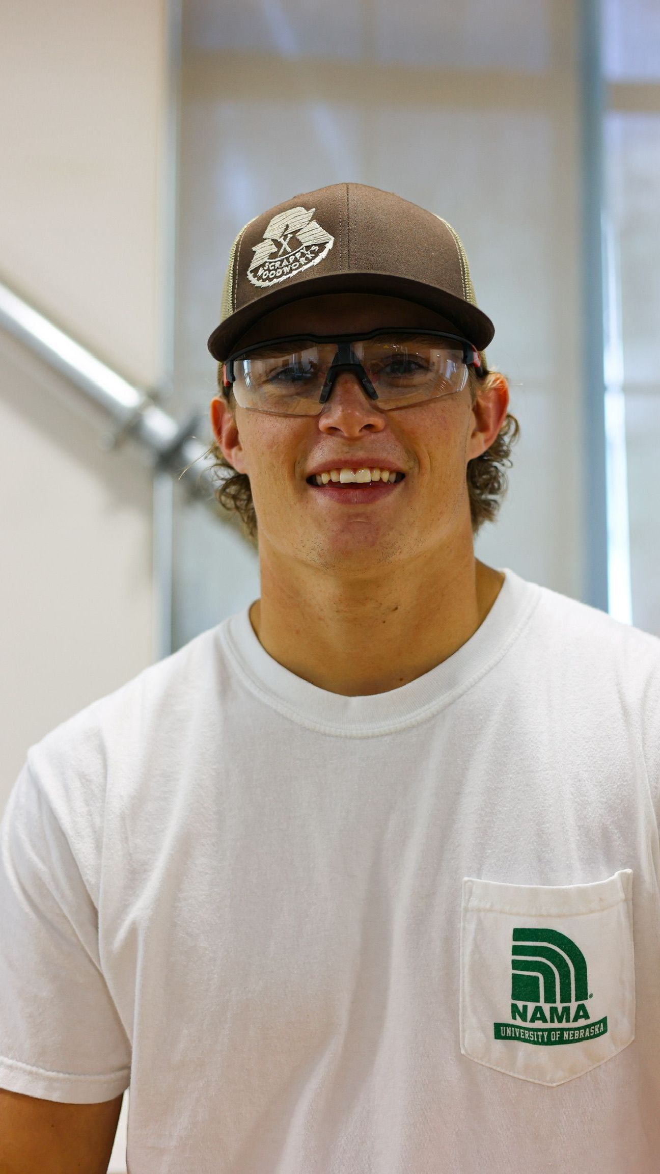 Man wearing a hat and safety glasses, smiling at the camera. He has a white shirt with a pocket, set indoors.