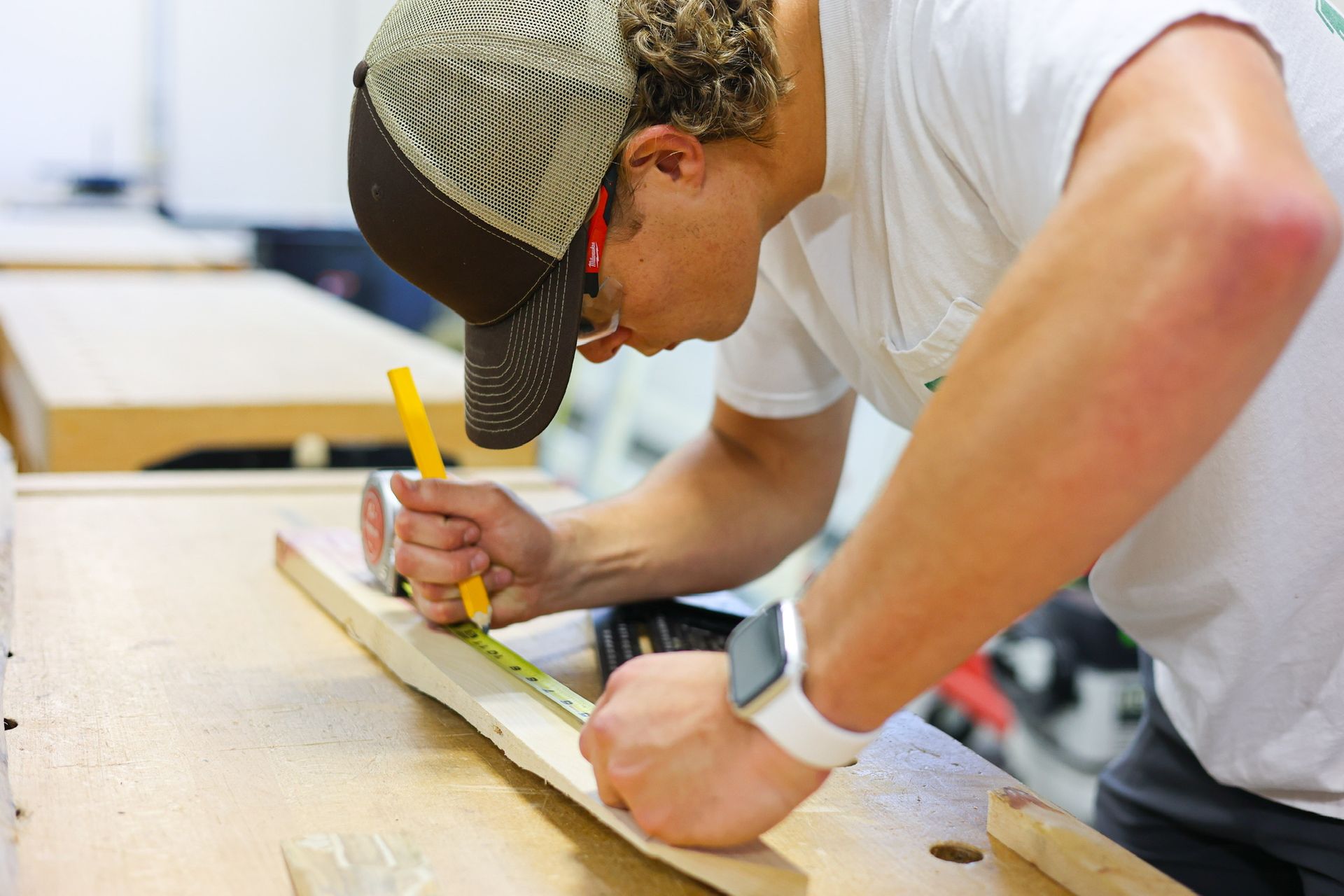 A man in a cap measures and marks a wood plank with a pencil and measuring tape at a workbench.