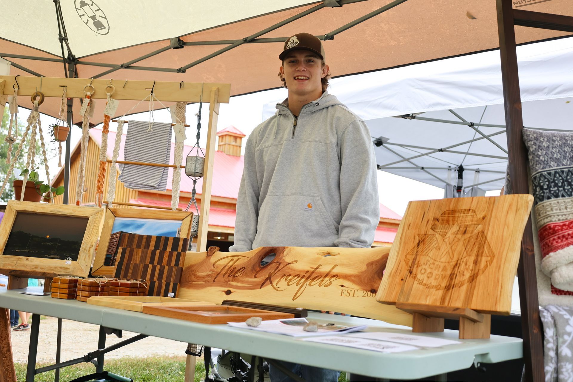 A young man stands behind a table displaying wooden crafts at an outdoor market.