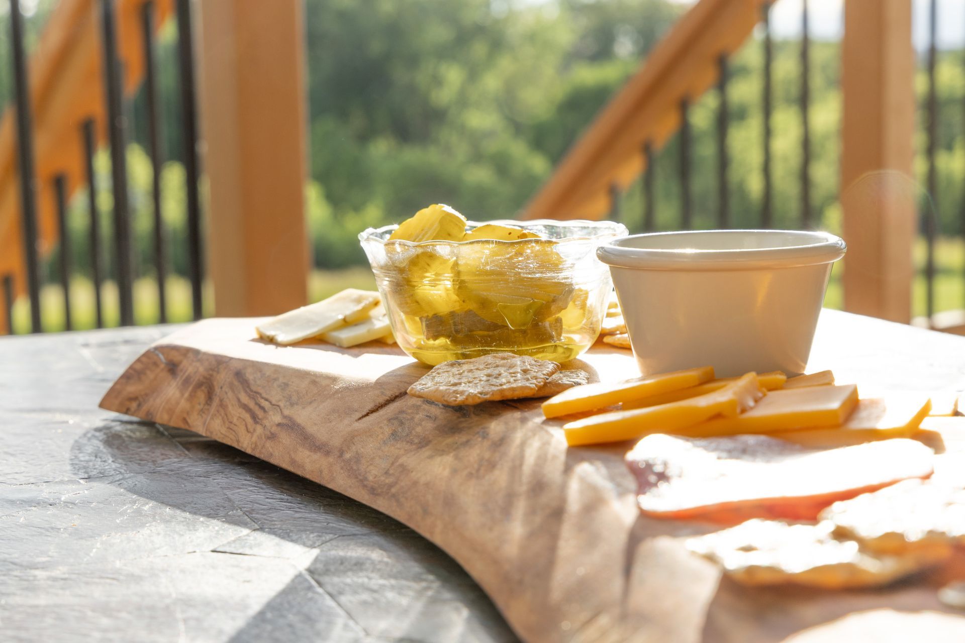 Wooden board with crackers, cheese, and a dip, set on a table on a deck with greenery in the background.