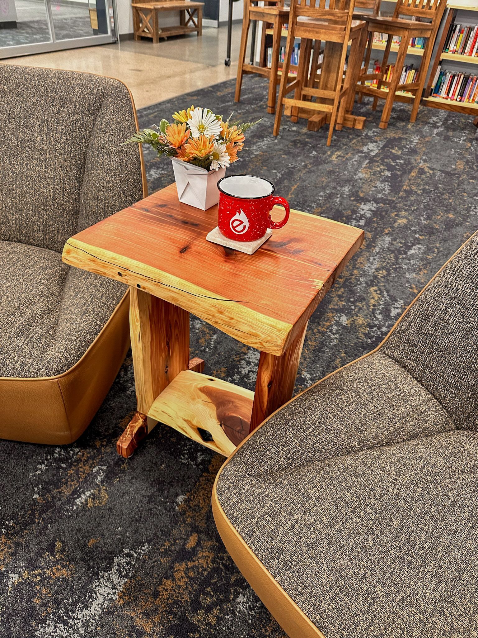 Wooden table with mug and flowers between two speckled armchairs; retail setting.