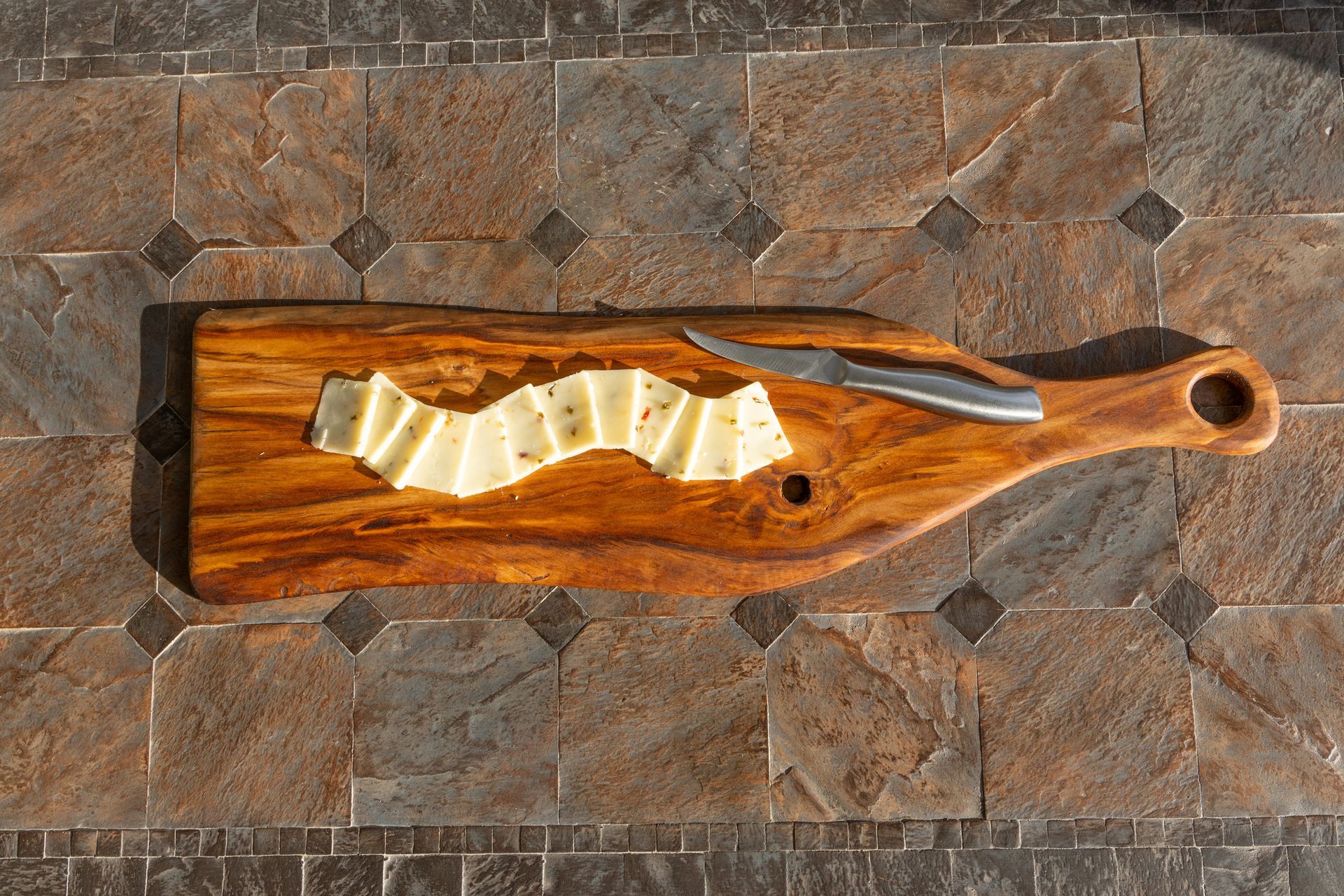 Wooden board with sliced cheese and a knife on a stone-tiled surface.