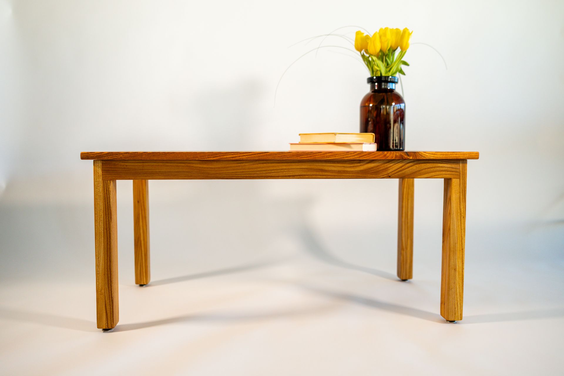 Wooden coffee table with amber vase of yellow flowers and two books on a white background.