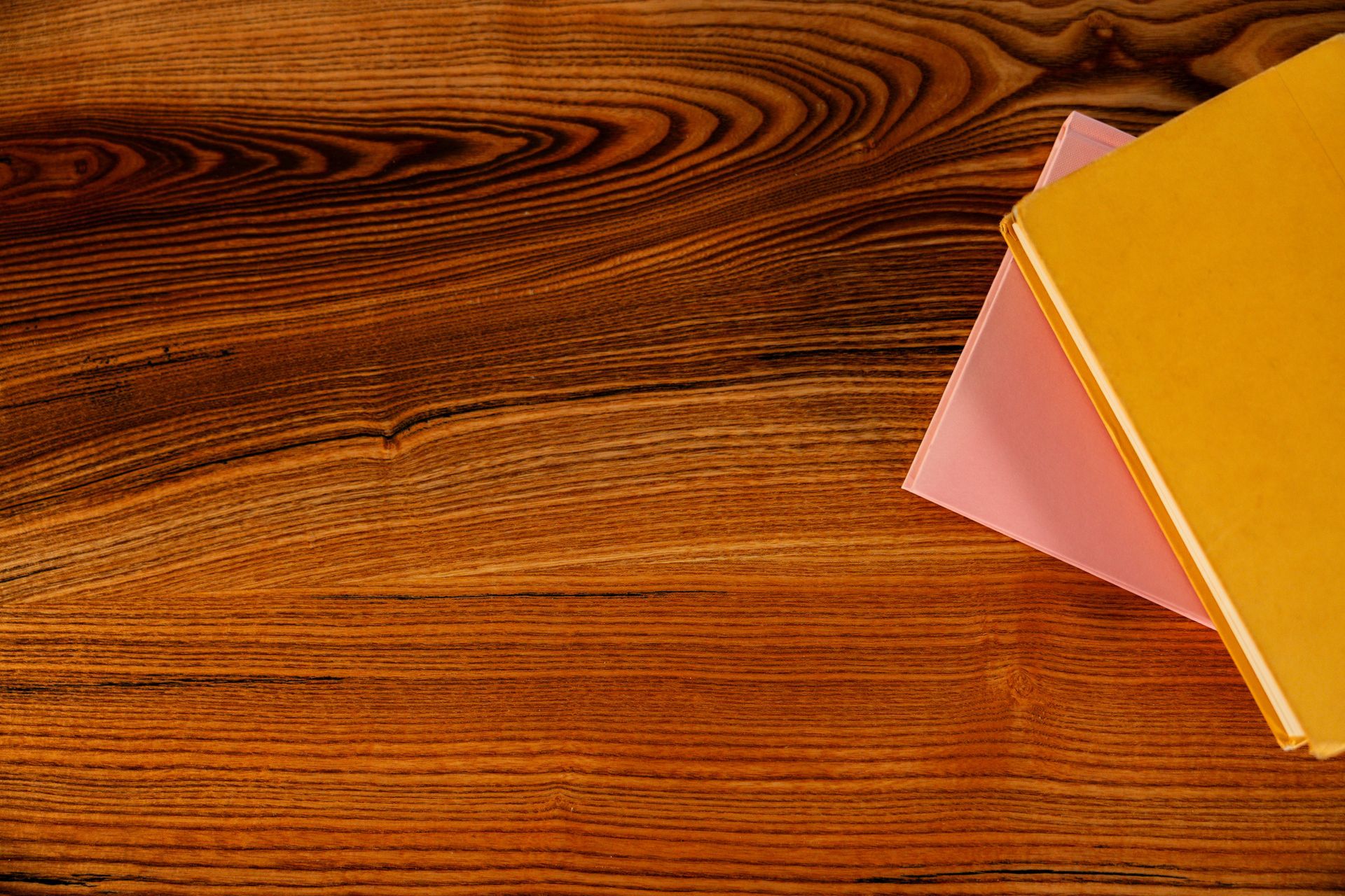 Books stacked on a wooden table: a yellow book on top of a pink one.