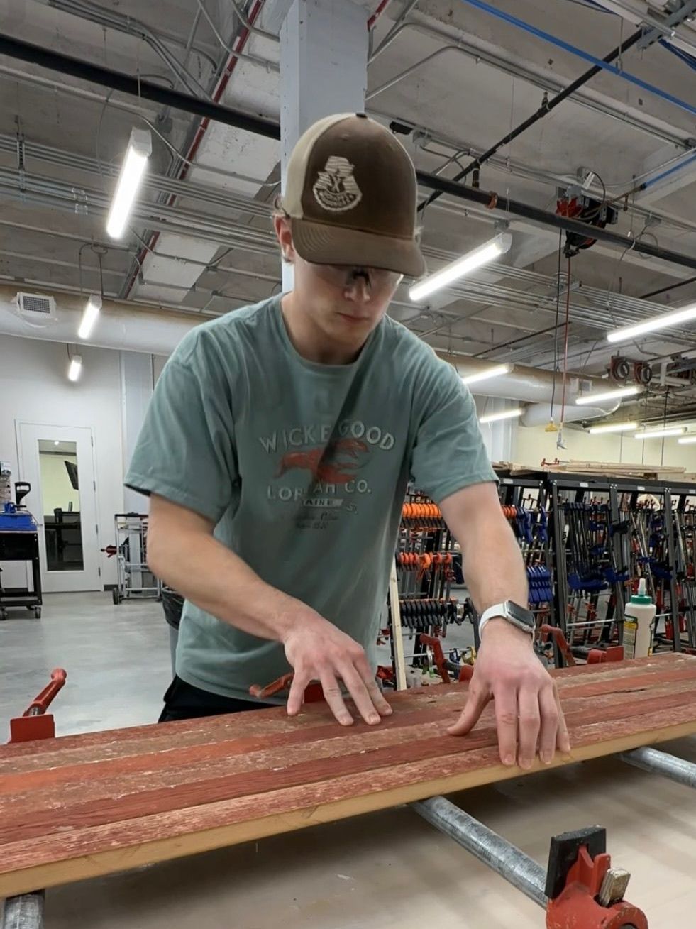 Man in workshop wearing hat, glasses, working with wood, hands pressed down, clamped in place.