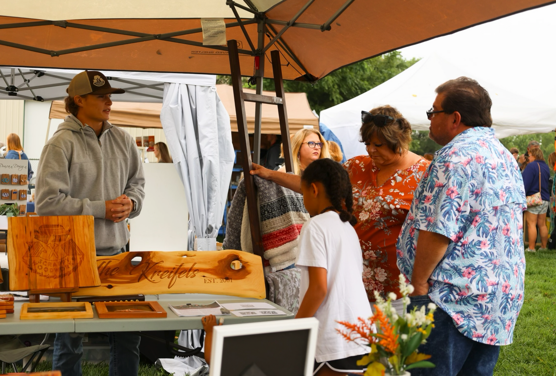 People browsing wood crafts at an outdoor market; vendor stands under tents; sunny day.