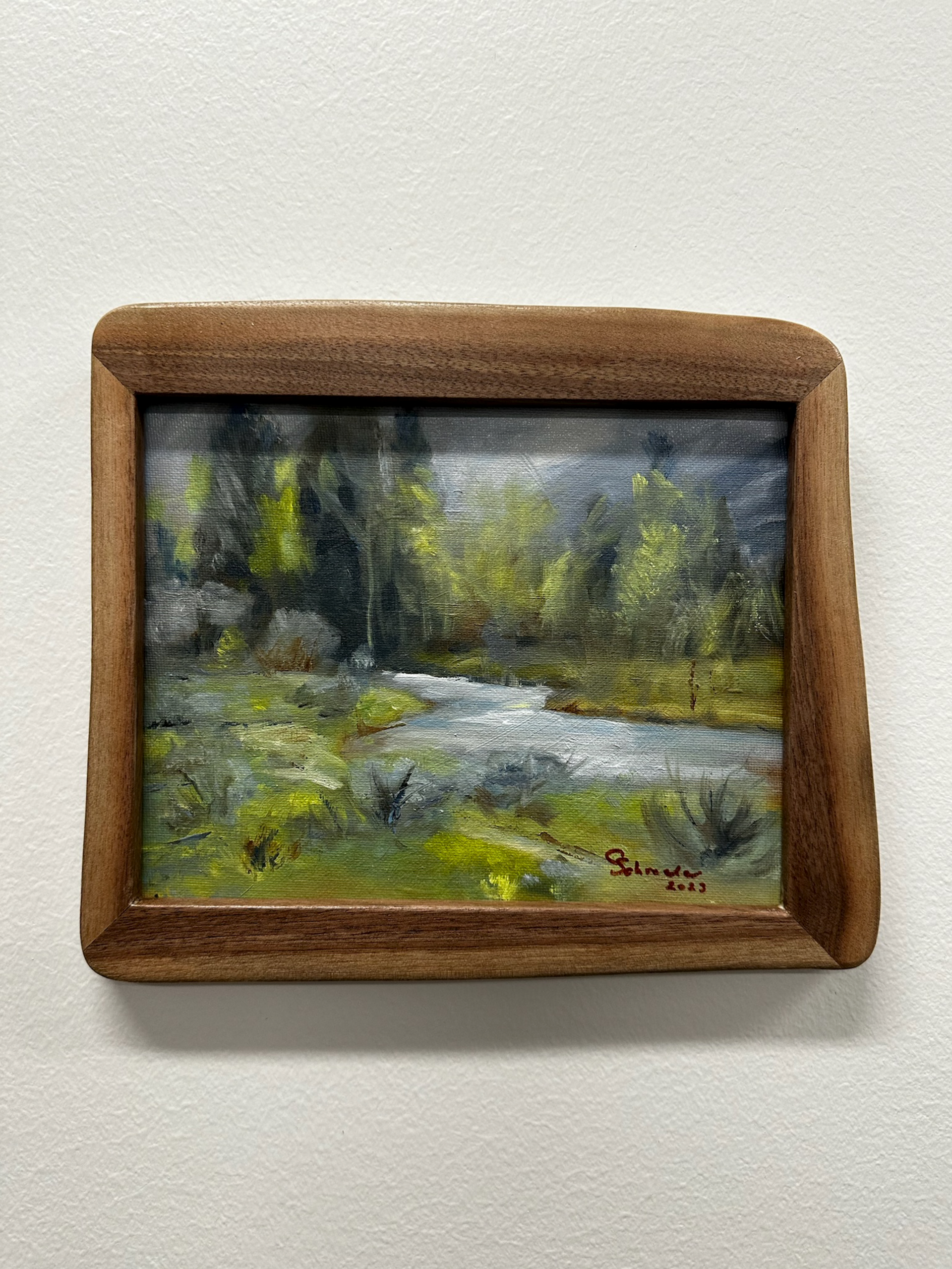 Painting of a river flowing through a forest, in a brown wooden frame.