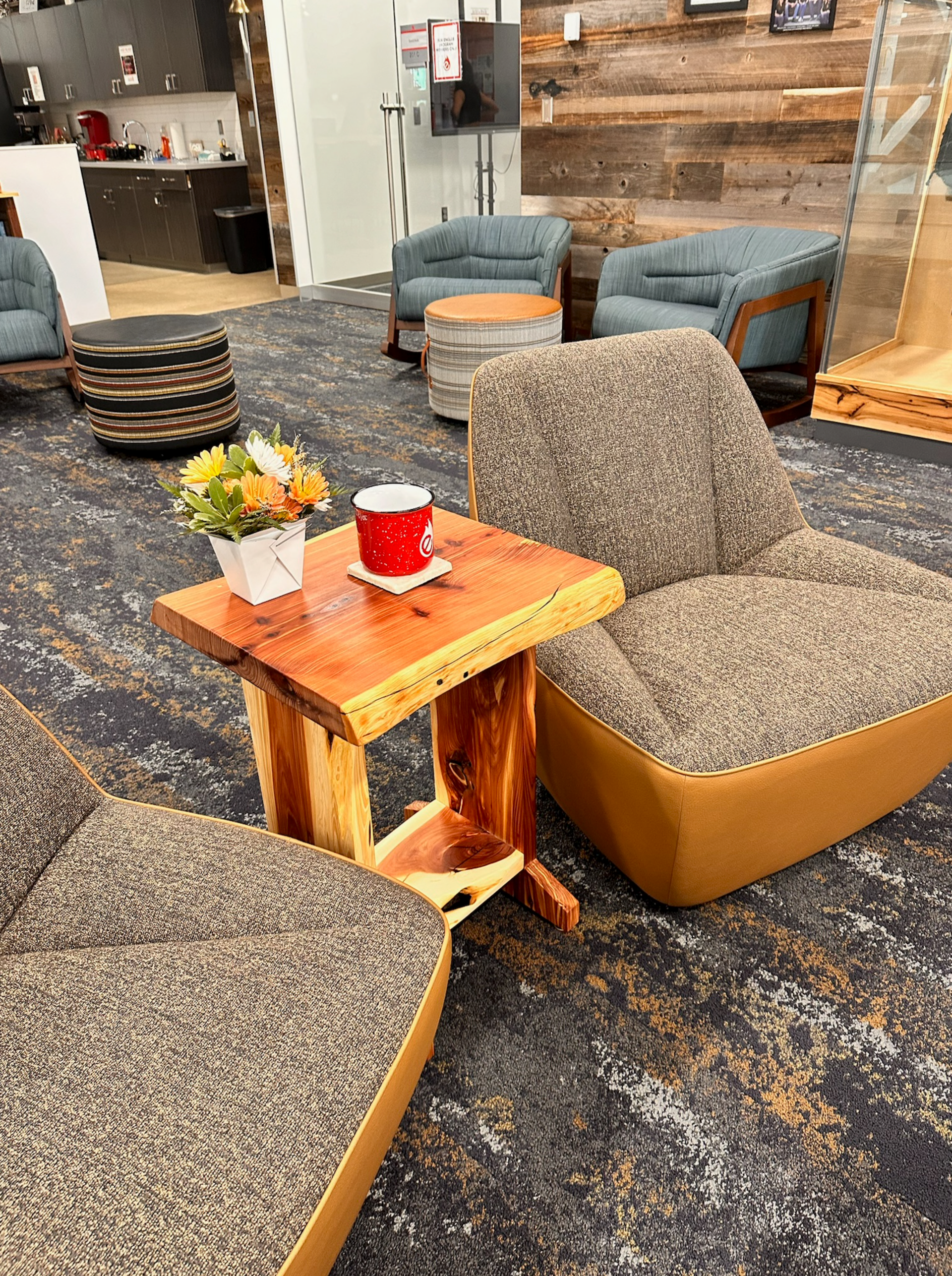 Rustic wooden coffee table with a red mug and flowers, surrounded by lounge chairs in a modern office setting.