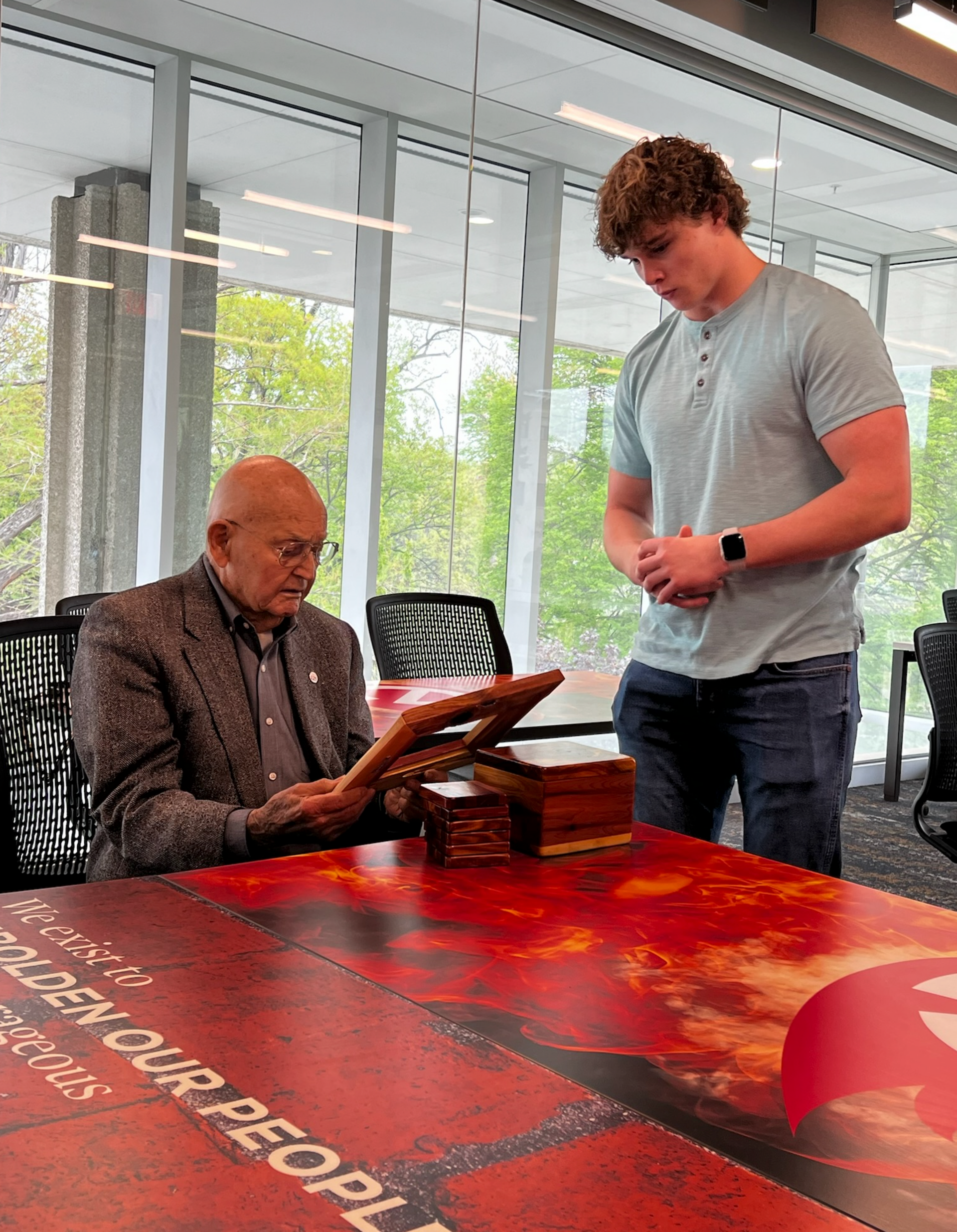 An older man reads a book at a table as a young man looks on in an office.