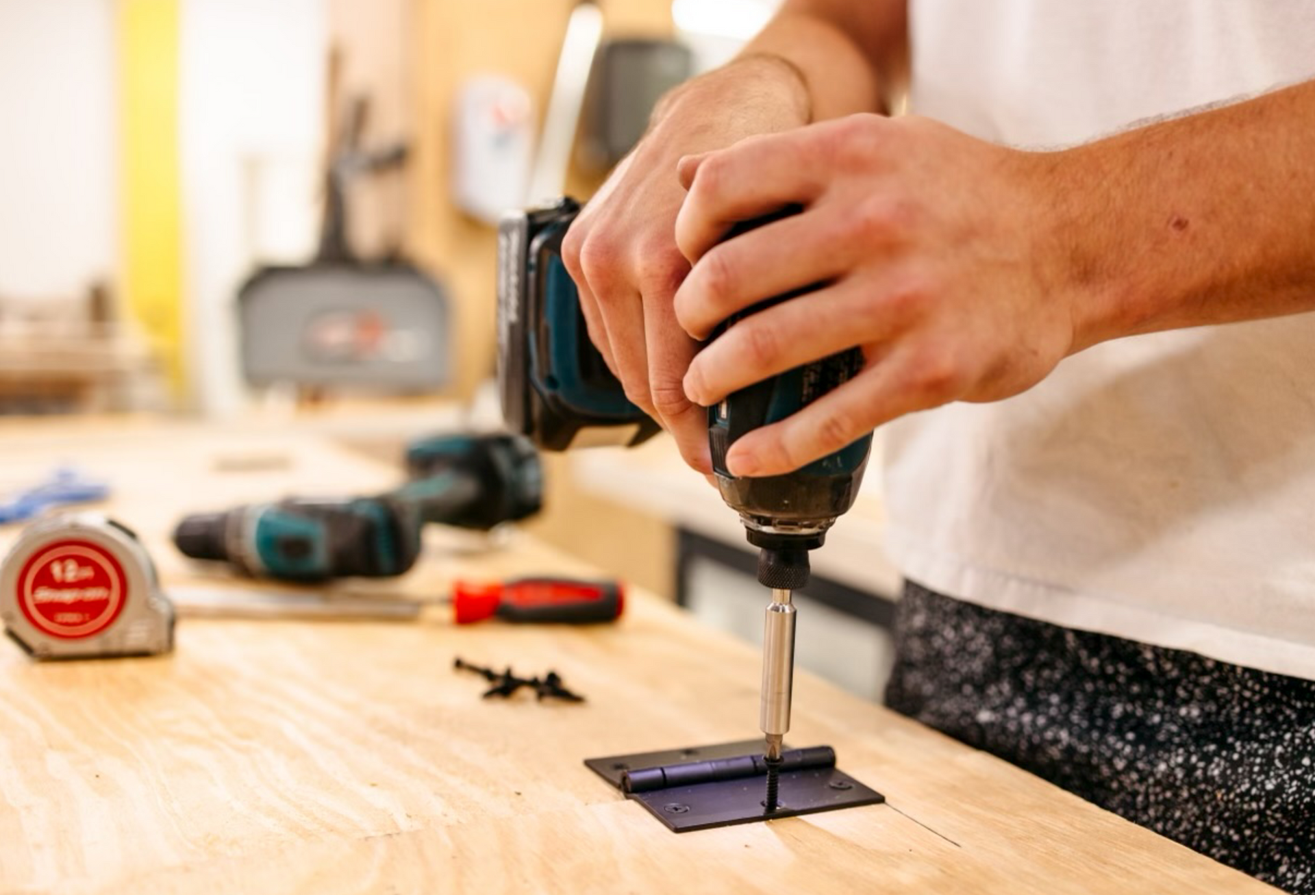 Person using a power drill to attach a hinge to a wooden surface in a workshop.