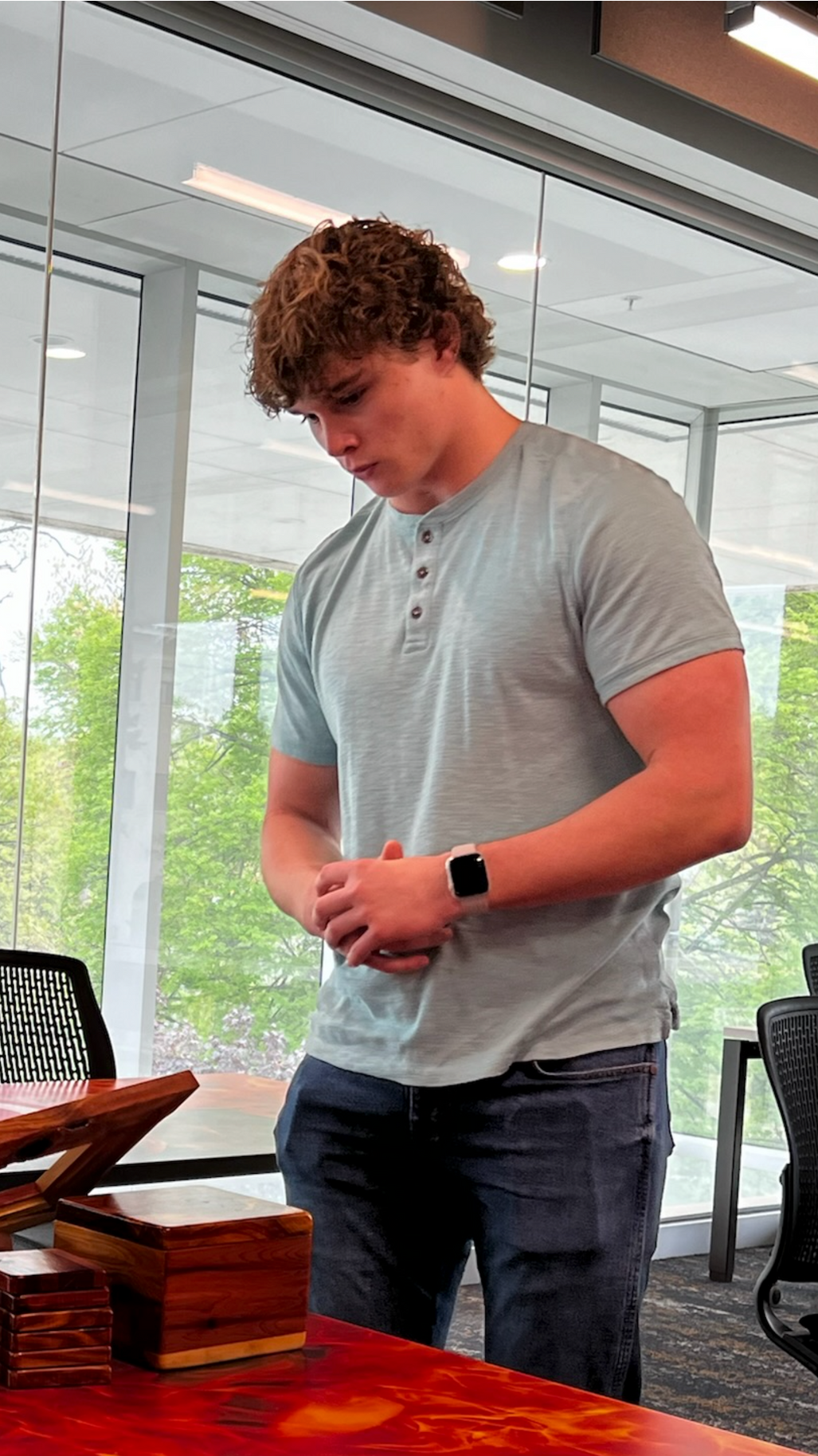 Man with curly hair in a gray shirt and jeans stands near a desk in a well-lit room.