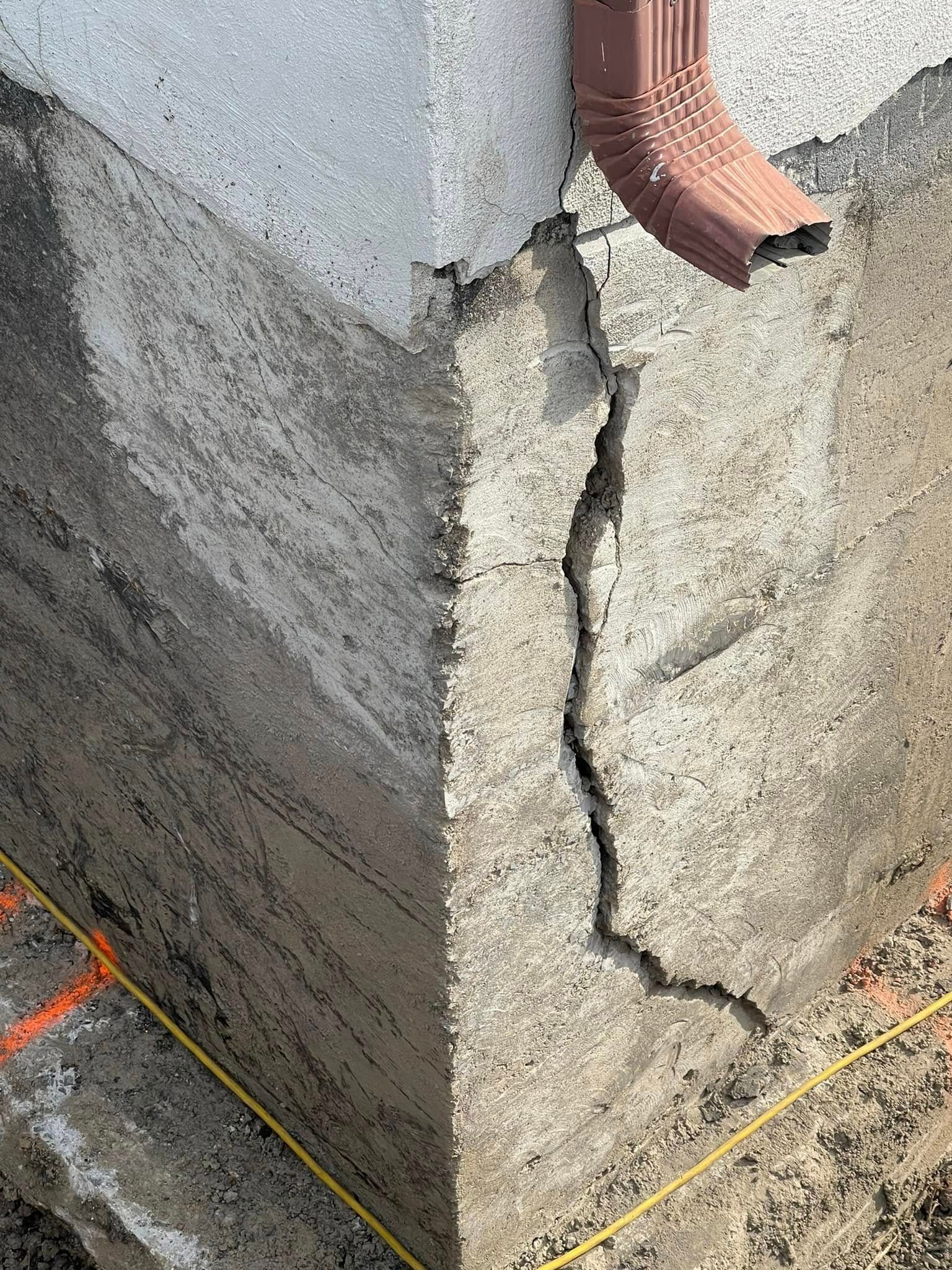 Coin de mur en béton fissuré avec une fente verticale visible et un tuyau de drainage rouge au-dessus.
