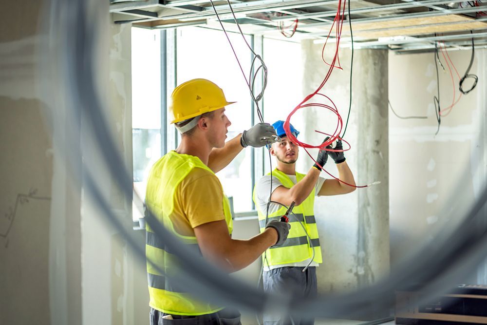 Two electricians working on wiring in a construction site, wearing safety vests and hard hats.