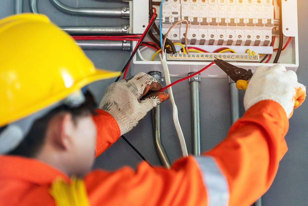 Electrician in orange overalls and yellow hard hat, wiring a circuit box.